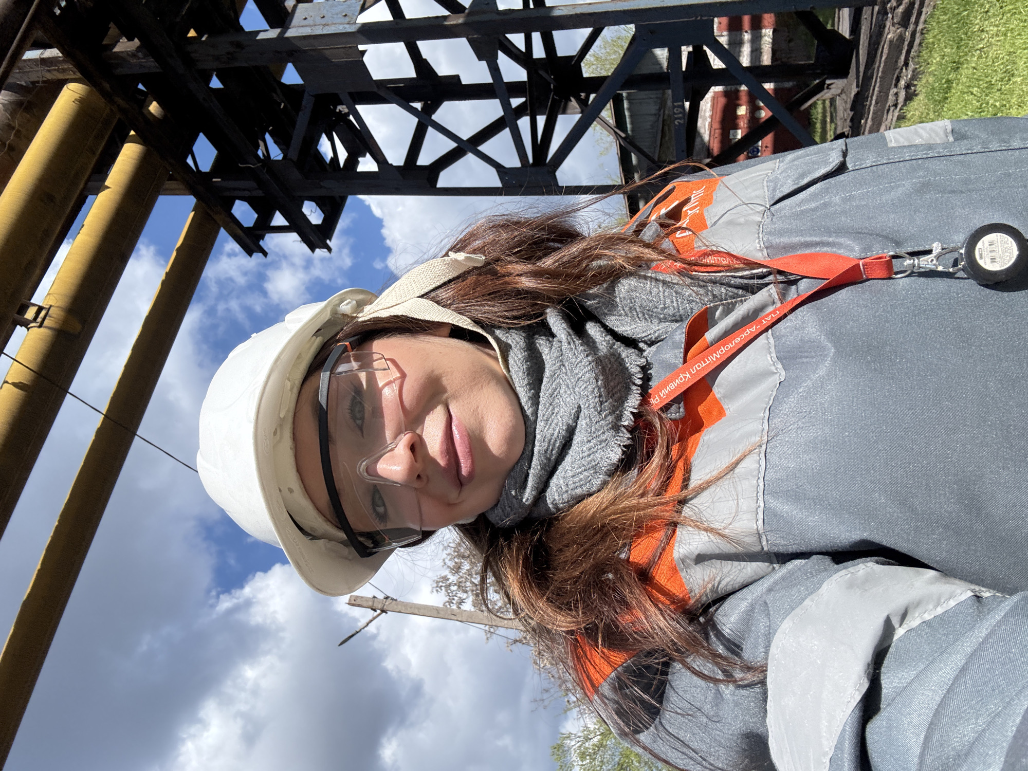 A medium shot depicts a woman wearing a white hard hat and safety glasses, with a gray scarf around her neck. She is wearing a gray work jacket with orange reflective stripes. An orange lanyard with a retractable badge holder is visible around her neck. She appears to be in an industrial area, as evidenced by the steel girders and yellow pipes in the background. The sky is visible with scattered clouds. Based on the lanyard inscription "ПАТ АРСЕЛОРМІТТАЛ КРИВИЙ РІГ," the location is likely related to ArcelorMittal Kryvyi Rih, a steel plant in Kryvyi Rih, Ukraine.