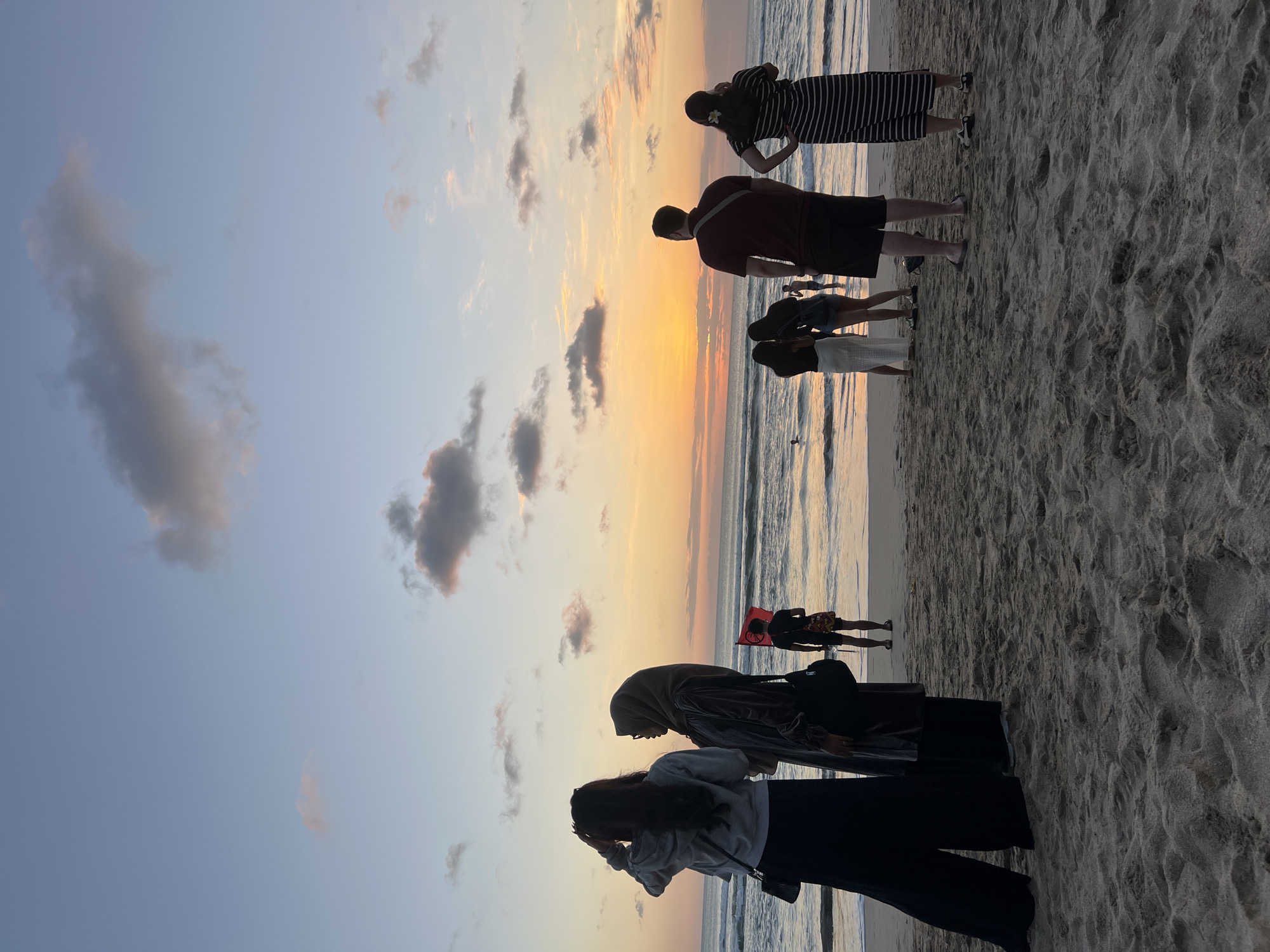 The image depicts a serene sunset scene on Kuta Beach, Indonesia. Silhouettes of people are visible on the sandy shore, with the ocean waves gently rolling in the background. The sky is a canvas of soft hues, transitioning from pale blue to warm oranges and yellows as the sun dips below the horizon. A few scattered clouds add texture to the sky. In the mid-ground, a person in the distance holds up a red flag. The overall atmosphere is tranquil, suggesting a peaceful end to the day.