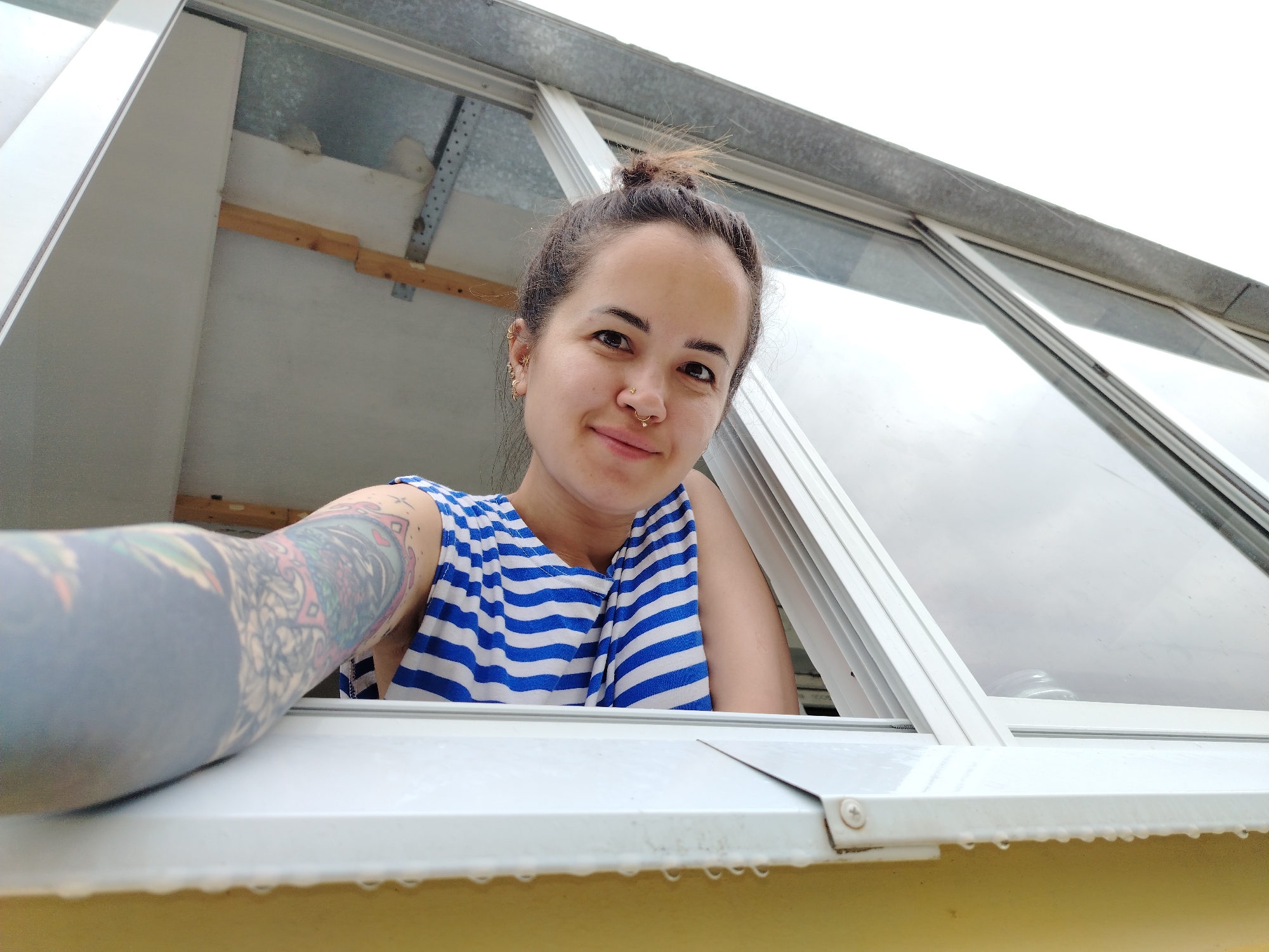 A young woman is captured in a close-up, low-angle shot, looking out of an open window from an interior space. She has dark hair tied in a messy bun, dark eyes, and a slight, pleasant smile. Multiple piercings adorn her ears, and she has a stud piercing on her nose and a hoop piercing in her septum. She wears a blue and white striped sleeveless top. Her left arm, heavily tattooed with a colorful and dark-shaded sleeve, rests prominently on the white window sill in the foreground.
The perspective is from below the window, looking up at the woman and the window opening. The window frame is white, and through the glass panes, an overcast or bright sky is visible. Inside the room, behind the woman, hints of a structural ceiling or upper wall can be seen, including wooden beams and metal supports, suggesting an attic, a conservatory, or a similar interior space.
Small water droplets are visible hanging from the underside of the white window sill, indicating recent rain or high humidity. The overall lighting is bright and natural, consistent with daytime. The woman's expression conveys a sense of calm or contentment. No text is visible in the image. The location context is Noviy, Russia.