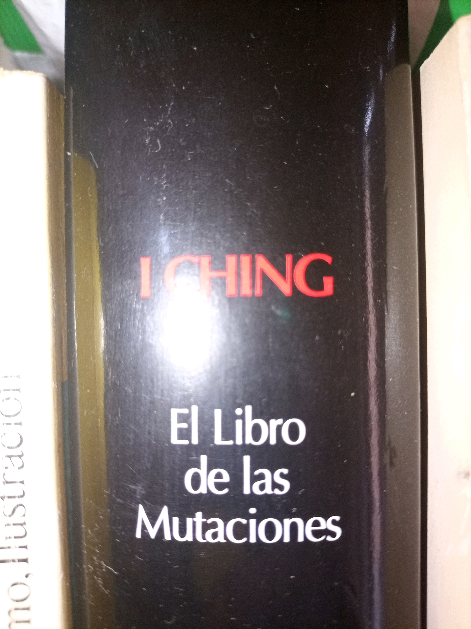 The image is a close-up shot of the spine of a book. The book has a black cover with red lettering that reads "I CHING" at the top. Below this, in white lettering, it reads "El Libro de las Mutaciones". The book is positioned vertically, with the spine facing the camera. To the left of the black-spined book, a portion of another book with a cream-colored spine is visible, with text that appears to be in Spanish. To the right of the black-spined book, another book with a white spine is partially visible. The lighting suggests an indoor environment, possibly a library or bookstore, with a bright light source creating a glare on the black book cover. There are no people or discernible city elements visible in the image. The location in Tarragona, Spain, cannot be determined from the provided image.