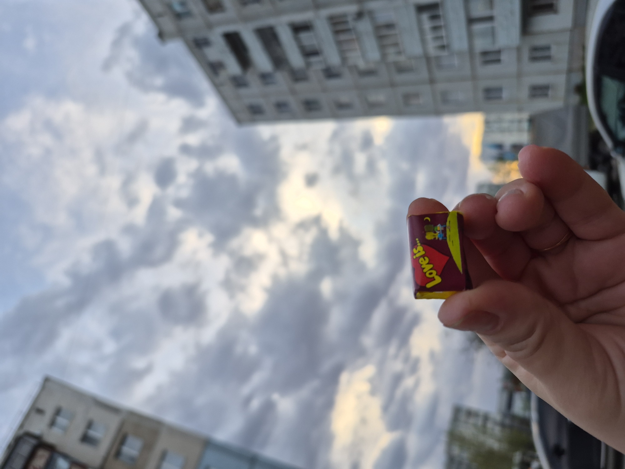 A person holds a wrapped Love is... chewing gum candy. In the background is an overcast sky and apartment buildings. The location is Tobolsk, Russia.