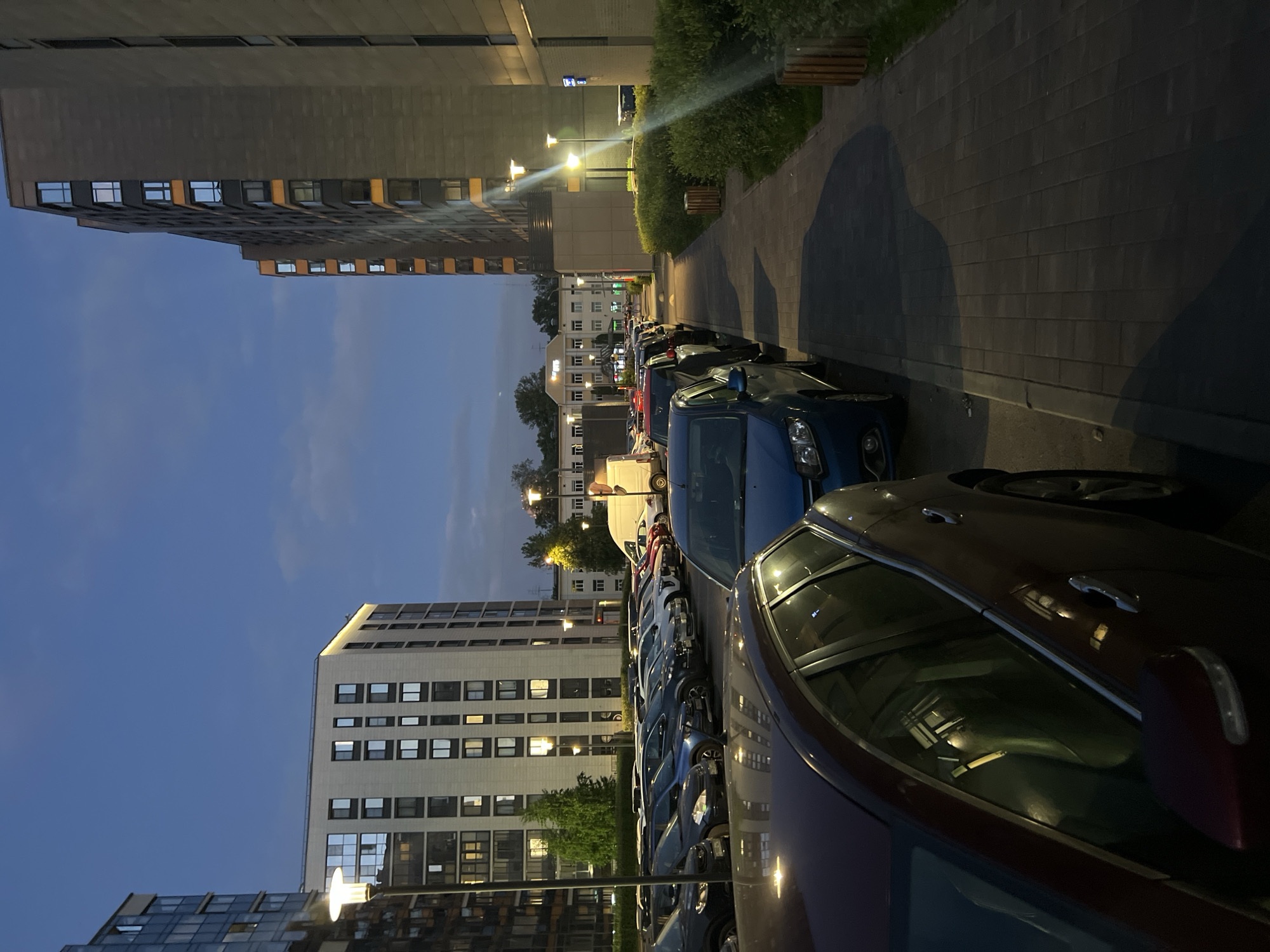 The image displays a street scene captured during late dusk or early night in Saint Petersburg, Russia, specifically identifiable by the "ГлавТрансПроект" (GlavTransProekt) building located at Obvodny Canal Embankment, 122, which is visible in the background.
The scene is dominated by modern architecture and an active parking area. In the immediate foreground on the left, the rear and side of a dark red station wagon are prominent. Directly to its left, the front of a blue sedan is visible. Beyond these, an asphalt parking lot extends into the midground, occupied by approximately two dozen parked passenger vehicles, including sedans and hatchbacks, in colors such as dark blue, black, red, and silver.
On the left side of the image, a multi-story residential building with a light-colored facade and numerous windows stands. Another similar light-colored residential building is situated in the center-left background, behind the parking lot. On the right, two tall, dark-grey residential buildings featuring subtle orange architectural accents are visible, with lights illuminated in many windows. In the distant center-background, the white "ГлавТрансПроект" building, displaying its illuminated Cyrillic sign and logo, is discernible. A white panel van is parked in front of this building.
A paved sidewalk, constructed from grey interlocking bricks, runs along the right side of the image, bordered by well-maintained green bushes. Several modern streetlights with upward-facing lamps are illuminated throughout the scene, casting light on the buildings, vehicles, and sidewalk. Two wooden trash bins are positioned alongside the sidewalk on the right. The sky is a deep blue with minimal cloud cover, indicating the transition from twilight to night. No human figures or discernible activities are present.