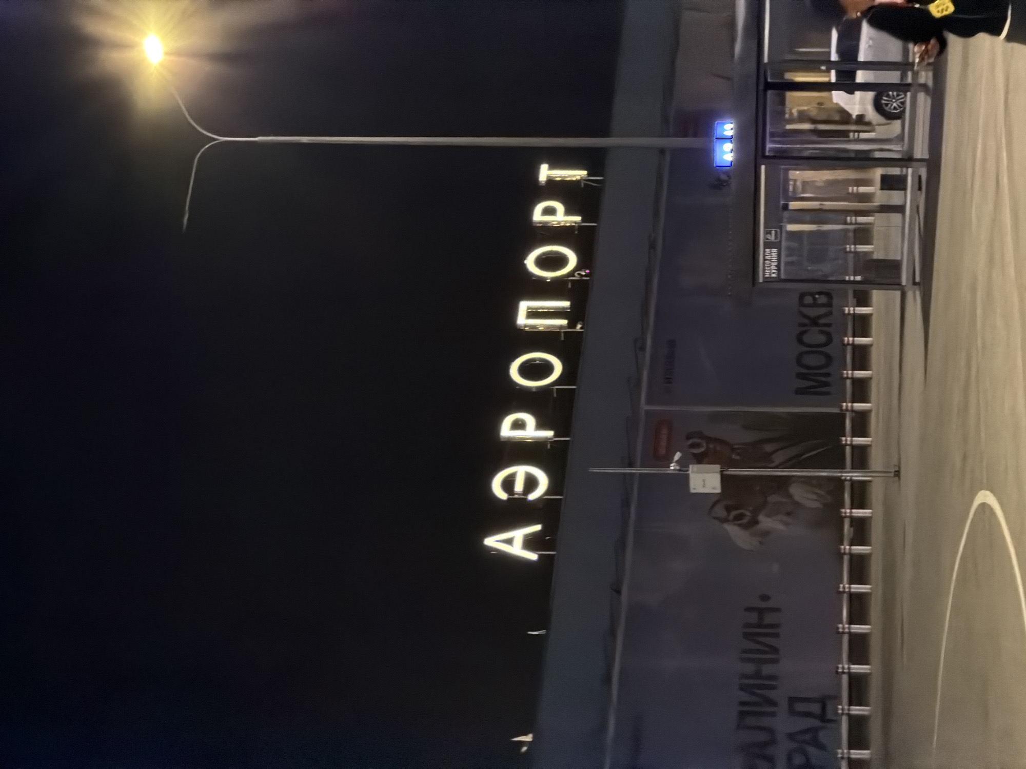 A night-time exterior shot shows the illuminated sign of an airport in Izhevsk, Russia, with the word "АЭРОПОРТ" (Aeroport) prominently displayed in large, bright letters. To the right of the sign, a tall street lamp with a bright light fixture is visible against the dark sky. In the foreground, a bus stop shelter with glass panels and a sign reading "МЕСТО ДЛЯ КУРЕНИЯ" (Smoking Area) is present. A white car is parked behind the bus stop. The ground in front of the bus stop appears to be a paved area, possibly a parking lot or roadway. A person in the lower right corner of the frame is walking away from the camera, holding a smartphone. A large advertisement banner is visible on the side of the building behind the bus stop, with text that includes what appears to be "КАЛИНИН...РАД" and "МОСКВ". The overall scene conveys an urban environment at night, likely at an airport terminal.