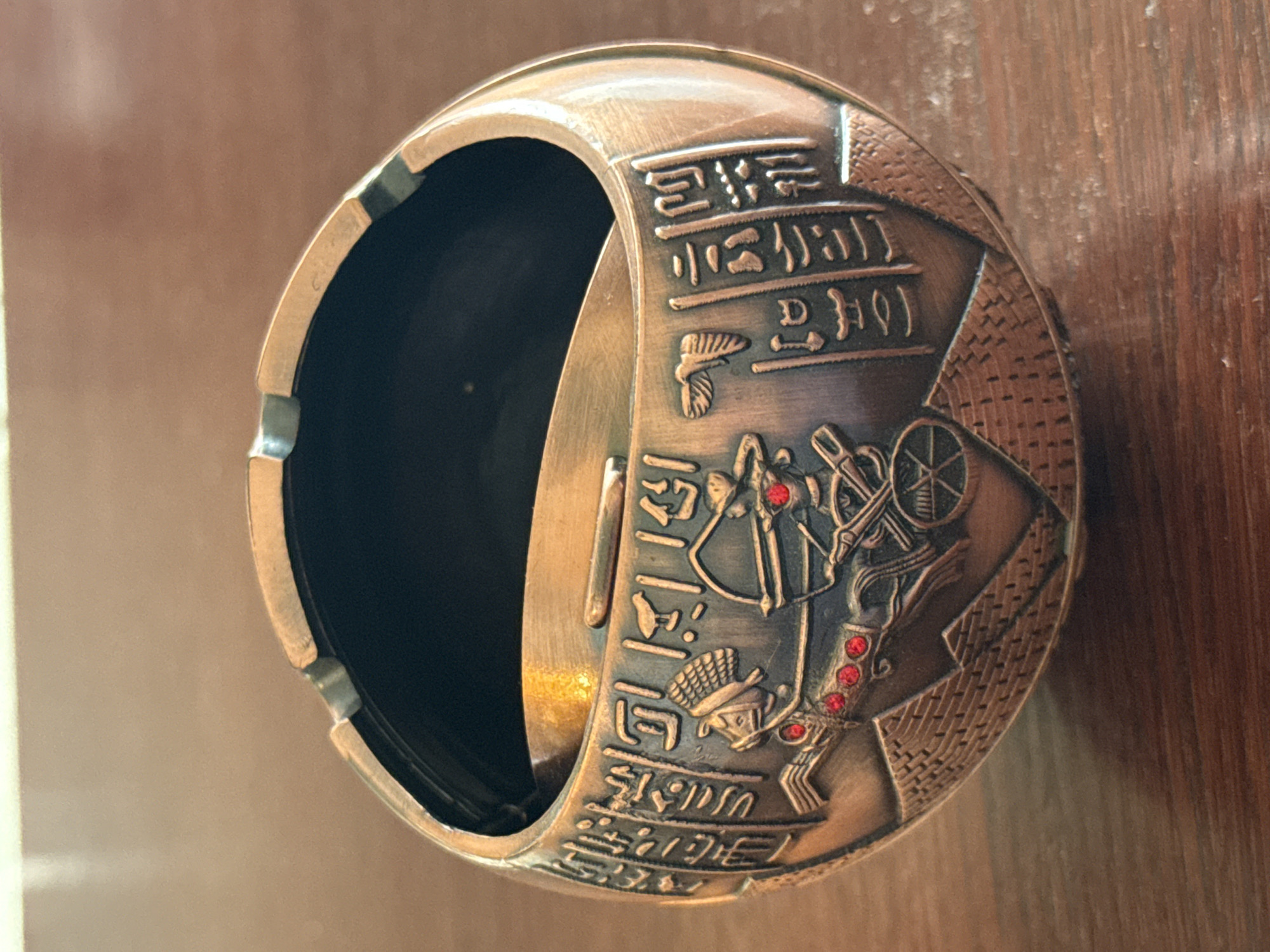 The media file shows a close-up, rotated view of a decorative, circular object with an opening, resembling a cuff or a bracelet. The object is made of a coppery-bronze colored metal and is intricately engraved with what appears to be Egyptian hieroglyphics and imagery.
The engraved imagery includes figures that suggest ancient Egyptian motifs, such as figures with pharaonic headdresses and depictions of chariots or elaborate machinery, possibly with wheels. There are also small, red, gem-like inlays embedded within the engravings, adding pops of color to the metallic surface. The hieroglyphics are arranged in rows, adding to the artifact's ancient aesthetic.
The object is placed on a surface that appears to be wood, with visible grain patterns, suggesting it might be a table or a shelf. The lighting suggests an indoor setting, and there are no discernible environmental cues like time of day or weather. The overall impression is that of an ornamental piece, possibly a collectible or a piece of jewelry, inspired by ancient Egyptian art and culture. The focus is entirely on the object itself, with the background being softly blurred, drawing the viewer's attention to the detailed craftsmanship.