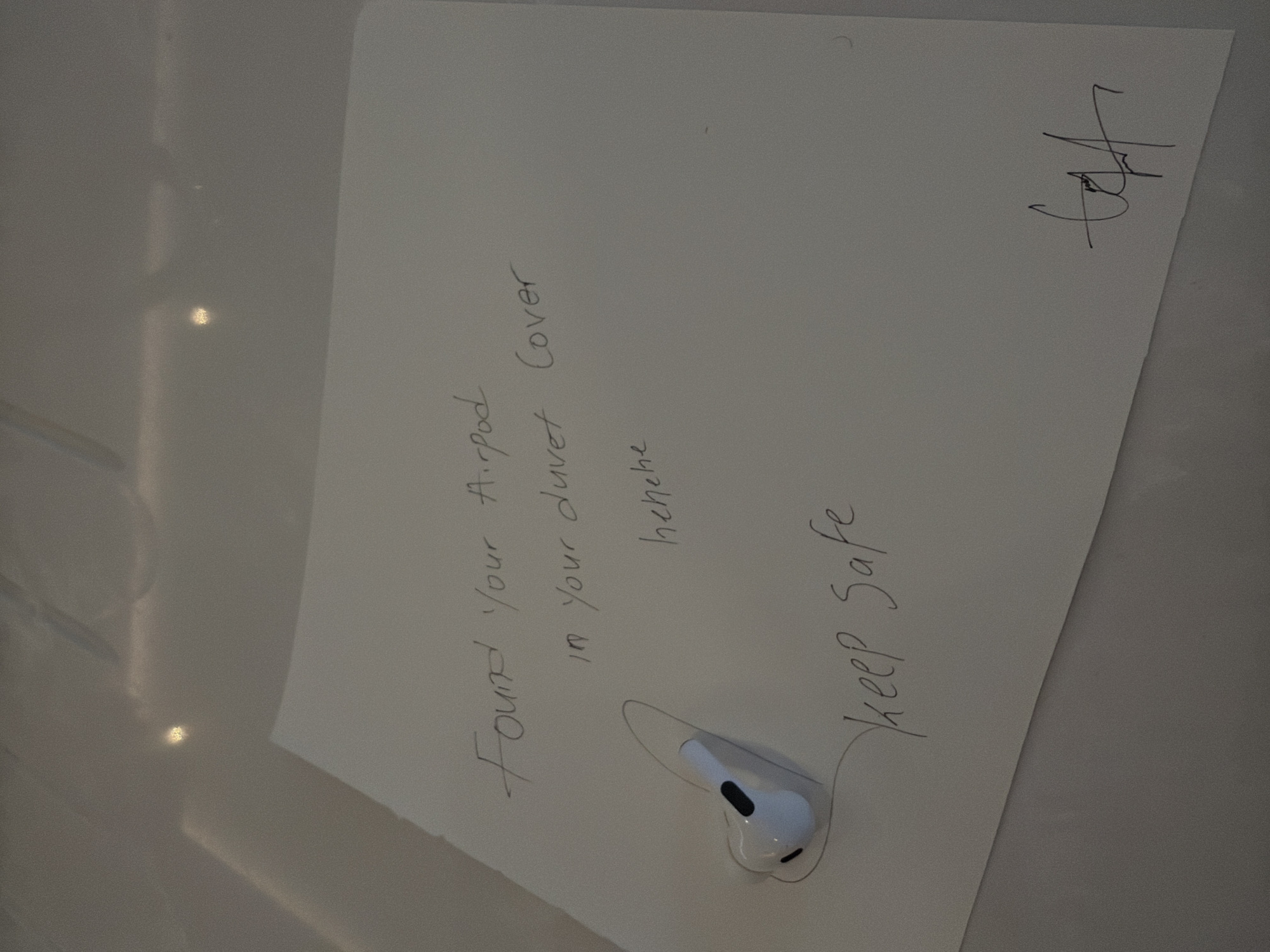 An AirPods Pro earbud is placed on a piece of paper. The paper has handwritten text. The top line reads "Found your Airpod". The second line reads "in your duvet cover". The third line reads "hehehe". The bottom left of the paper has a handwritten phrase "keep Safe". In the bottom right corner of the paper is a signature. The background appears to be a light-colored, reflective surface, possibly a table or desk. The lighting suggests an indoor environment. The image does not contain any information about the location in the city of London, United Kingdom.