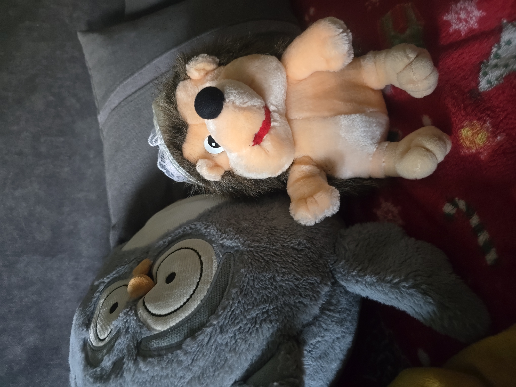 Two stuffed animals are on a red blanket with a candy cane pattern. The stuffed animals are placed on a grey couch. On the left is a large grey stuffed owl with white and black eyes and an orange beak. On the right is a smaller stuffed hedgehog with brown fur, a black nose, and a white and lace hat. The hedgehog has its arms outstretched and its mouth is open in a smile. The owl's eyes are wide open. A yellow object is partially visible on the left side of the image. The scene appears to be indoors, possibly in a residential setting. There is no indication of the city of Golyanovtsi, Bulgaria.