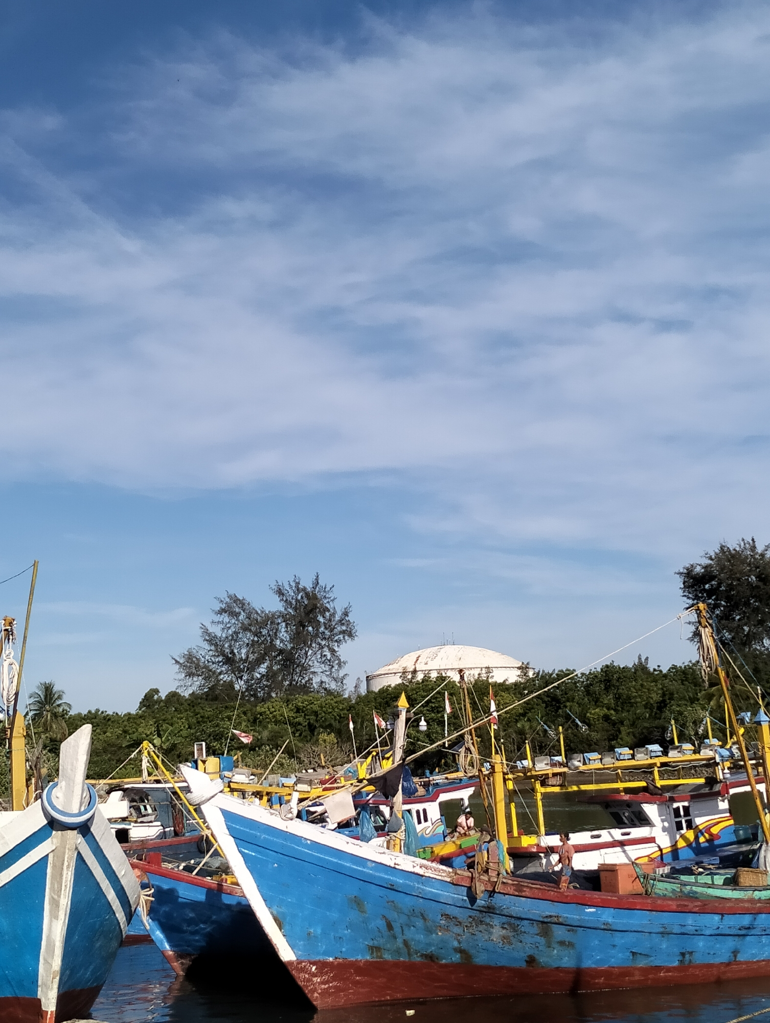 The image captures a harbor scene in Lhokseumawe, Indonesia, featuring several blue and white fishing boats docked in a harbor. The boats are traditional, with brightly colored hulls and intricate details. Two men are visible on one of the boats. The backdrop includes lush green vegetation and a large, white spherical structure on the horizon, potentially a water tank. The sky above is a light blue with scattered clouds.