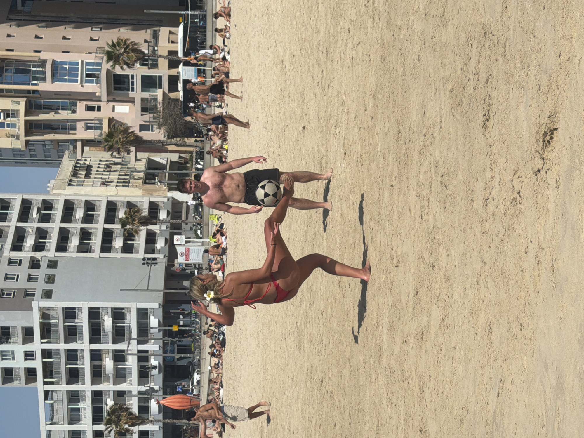 The image shows a crowded beach in Tel Aviv, Israel, with several people playing with a soccer ball. A man is preparing to kick the ball to a woman performing a bicycle kick. In the background, there are several high-rise buildings.