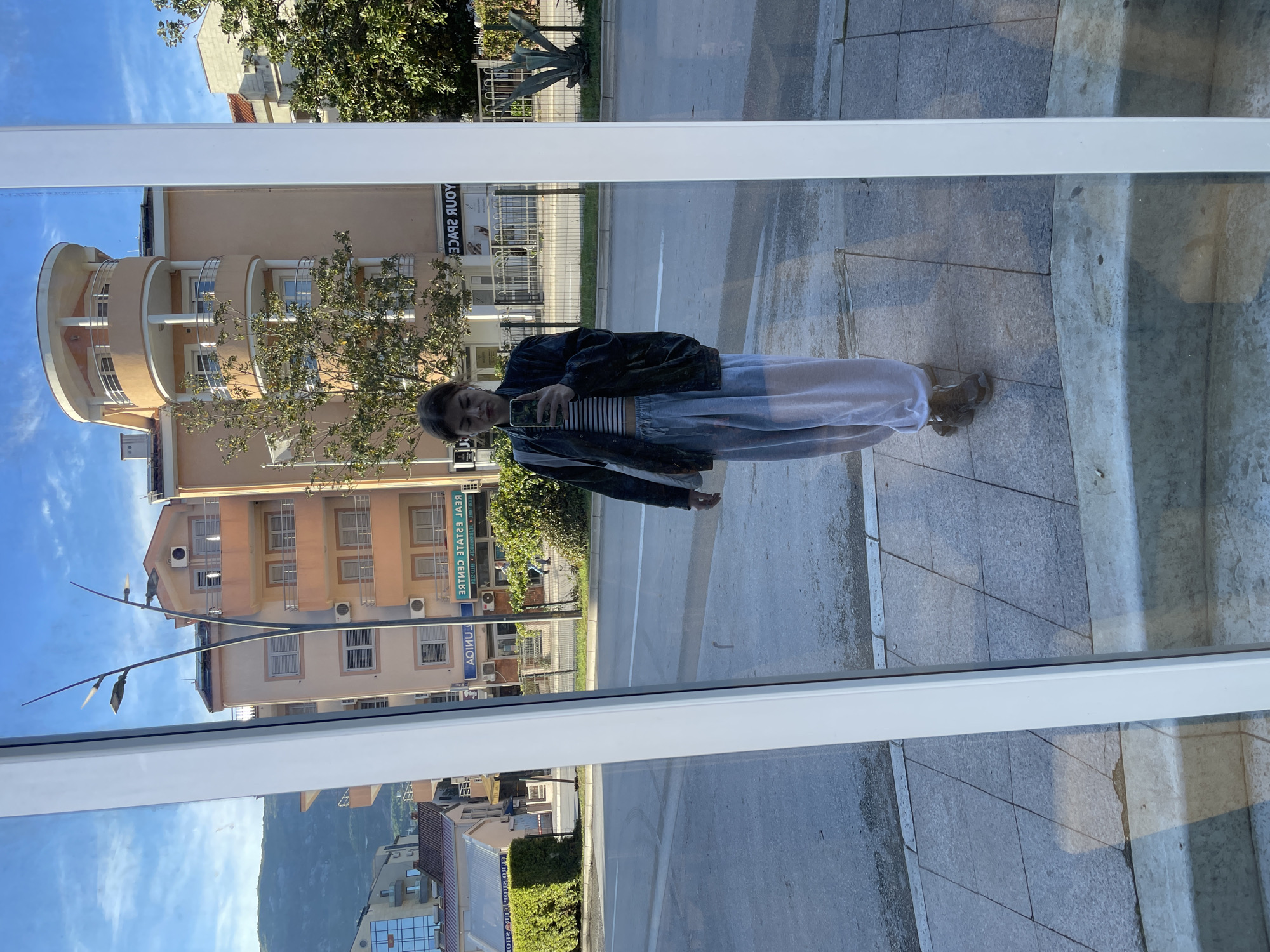 This image captures a reflection of a young woman taking a selfie in a large window or glass panel, set against an urban backdrop with natural elements. The scene is rotated 90 degrees clockwise from its natural orientation.
The primary subject is a young woman, seen in reflection, holding a smartphone to capture her image. She is wearing a dark jacket over a striped crop top and light-colored, possibly white or light blue, wide-leg pants or a skirt. Her expression is neutral, and her dark hair is pulled back.
The reflection reveals a vibrant outdoor setting. Behind the woman, a street or road is visible, flanked by sidewalks. On the opposite side of the street, there are several multi-story buildings under a clear blue sky. One prominent building to the left (when correctly oriented) features a distinctive curved facade with multiple balconies. Further down, other buildings with numerous windows and balconies are visible. In the distant background, mountains rise under the bright sky, characteristic of the Budva, Montenegro context.
Green trees and bushes are interspersed throughout the scene, with one crop showing a succulent plant (possibly an agave). The ground in the foreground, where the person is standing, is paved with square stone tiles. Streetlights with multiple arms are also reflected.
Several notable details include visible text on the reflected buildings. The words "YOUR SPACE" appear reversed on one building. Another building has the text "REAL ESTATE CENTRE" and "АГЕНЦИЈА" (AGENCIJA - Agency), both reversed, indicating a commercial area.
The time of day is bright daylight, suggested by the clear blue sky and strong reflections. The overall atmosphere is casual and urban, with the scenic mountainous landscape adding a natural dimension. The entire image is presented as a reflection, which is evident from the reversed text and the slightly distorted perspective of the scene.