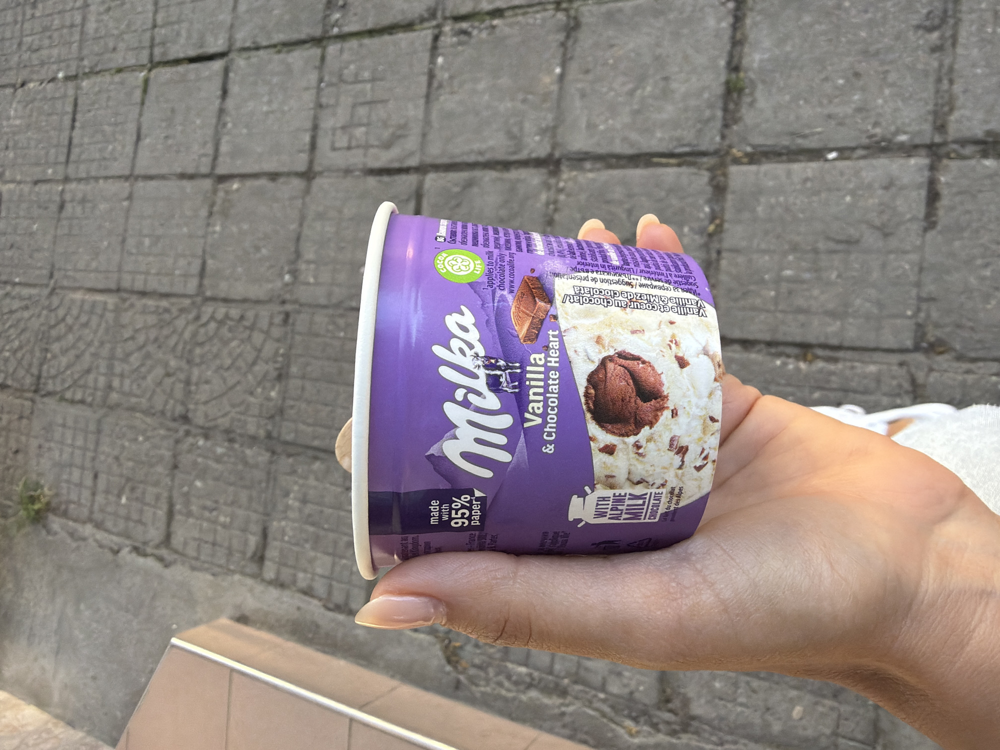 A person's hand, with manicured nails, holds a purple and white pint of Milka Vanilla & Chocolate Heart ice cream. The pint has an image of chocolate chunks and swirls of vanilla ice cream. The background is a gray paved walkway. The scene is outdoors.