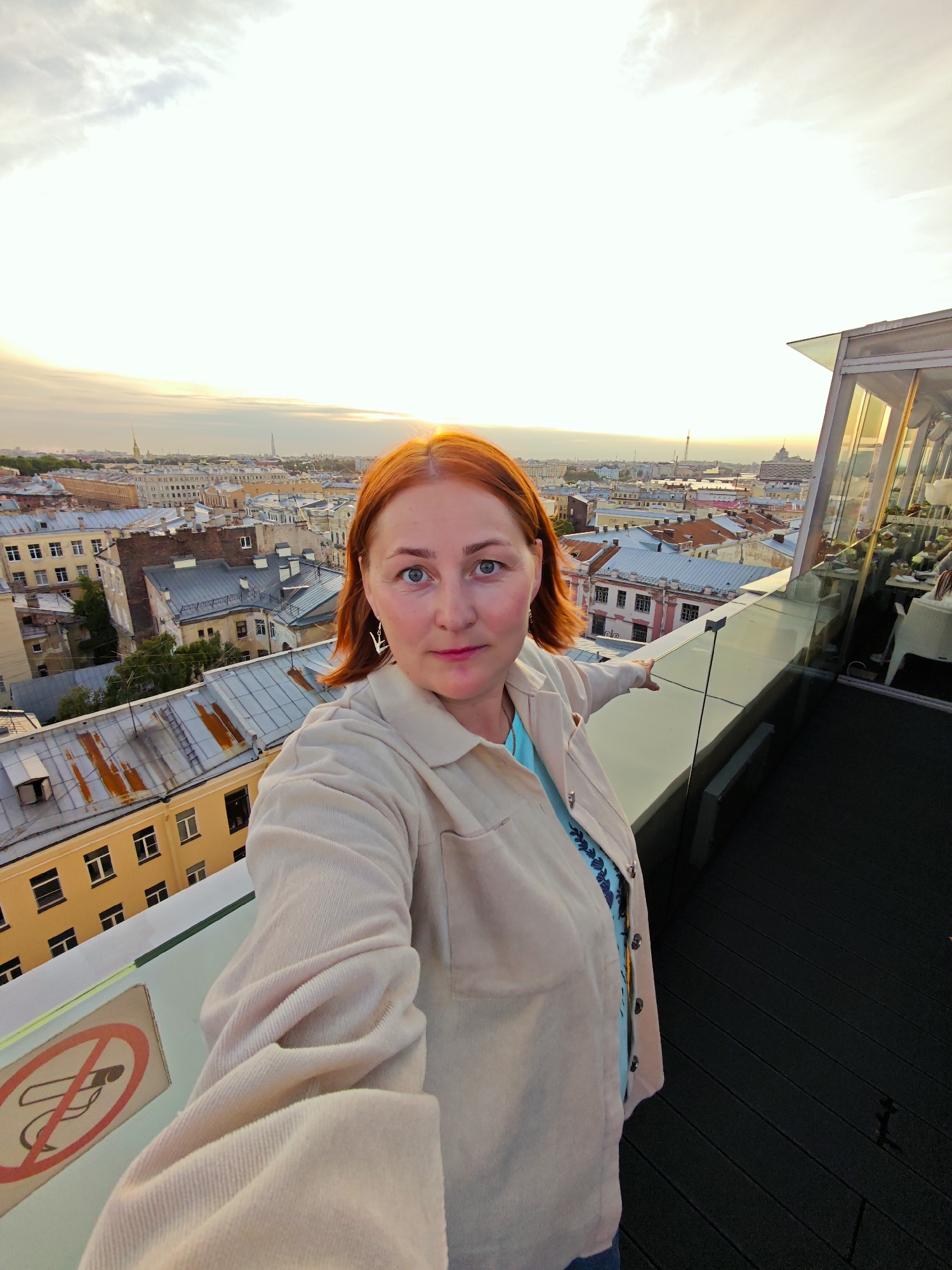 A woman with auburn hair is taking a selfie on a rooftop terrace overlooking the city of Saint Petersburg, Russia. She is wearing a beige corduroy jacket over a blue patterned top and a silver earring in the shape of a swan on her left ear. She is standing by a glass railing with a city skyline in the background under a pale, cloudy sky. The buildings below have terracotta tiled roofs, characteristic of Saint Petersburg's architecture. A "no smoking" sign is visible on the railing near her left arm. The terrace appears to be part of a restaurant or bar, with tables and chairs visible through the glass enclosure. The setting sun casts a warm glow on the cityscape.