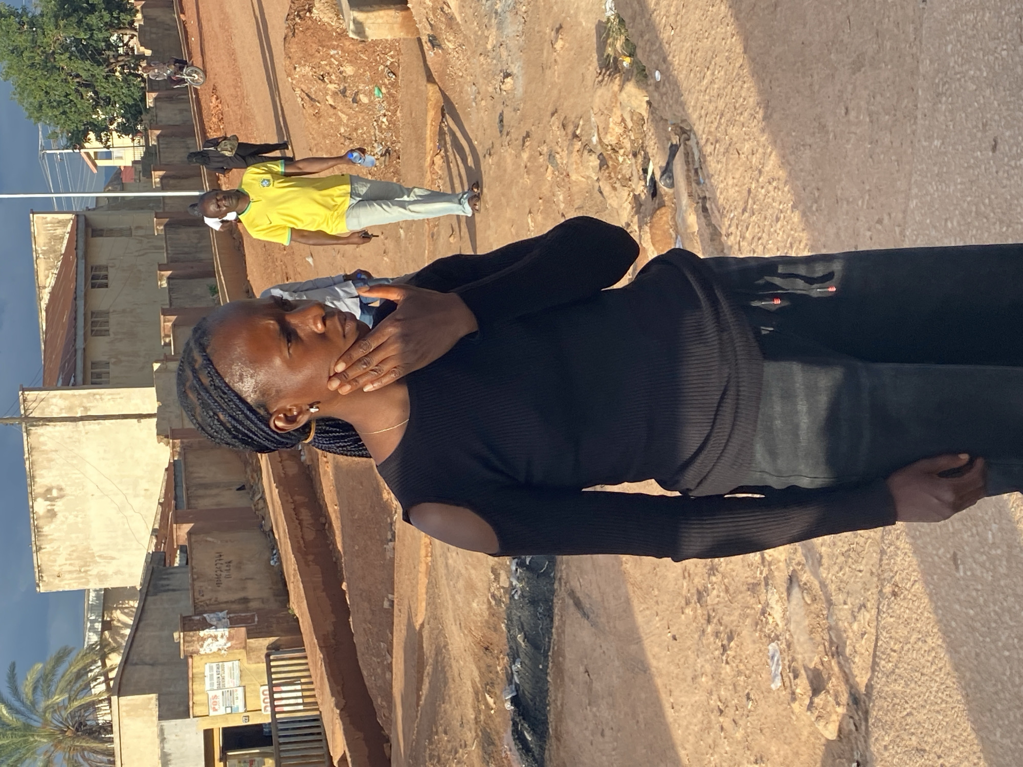 This image captures an outdoor street scene in Gura Top, Nigeria, during daylight hours. The primary subject is a Black woman in the foreground, positioned slightly to the left of the frame's center. She wears a black long-sleeved ribbed top with cold shoulder cutouts and dark pants. Her hair is braided, and she has a gold hoop earring in her left ear and a thin gold necklace. Her right hand is raised, touching her chin or cheek, and she gazes slightly to her right.
The ground is predominantly unpaved, composed of reddish-brown dirt with an uneven surface, showing tire tracks and footpaths. Scattered debris, including paper and plastic, is visible. In the middle ground, to the left of the main subject, there is a darker, wet-looking area containing accumulated refuse, suggesting a drainage ditch or collection point.
In the background, multiple individuals are present. A Black man, wearing a yellow t-shirt with a Brazil national football team emblem, light-colored pants, and flip-flops, walks towards the right side of the frame. He holds a clear plastic bottle in his right hand and a small dark object in his left. Behind him, another individual dressed in dark clothing, possibly with a head covering, walks away from the viewer, carrying a bag. Further back, a person rides a bicycle, and two additional figures are visible on a distant, elevated dirt path on the right.
The environment features several buildings of varying sizes. On the left, a light-colored building with a dark roof stands behind a concrete wall that displays weathered signs, including one with "POS" and another with the number "08162627741". Additional single and two-story structures, mostly constructed with light-colored plaster or concrete and some with corrugated metal roofs, populate the background. Vegetation includes a prominent palm tree on the far left and a leafy green tree on the far right. The sky is partially cloudy, and the lighting suggests late afternoon, with noticeable shadows cast across the ground.