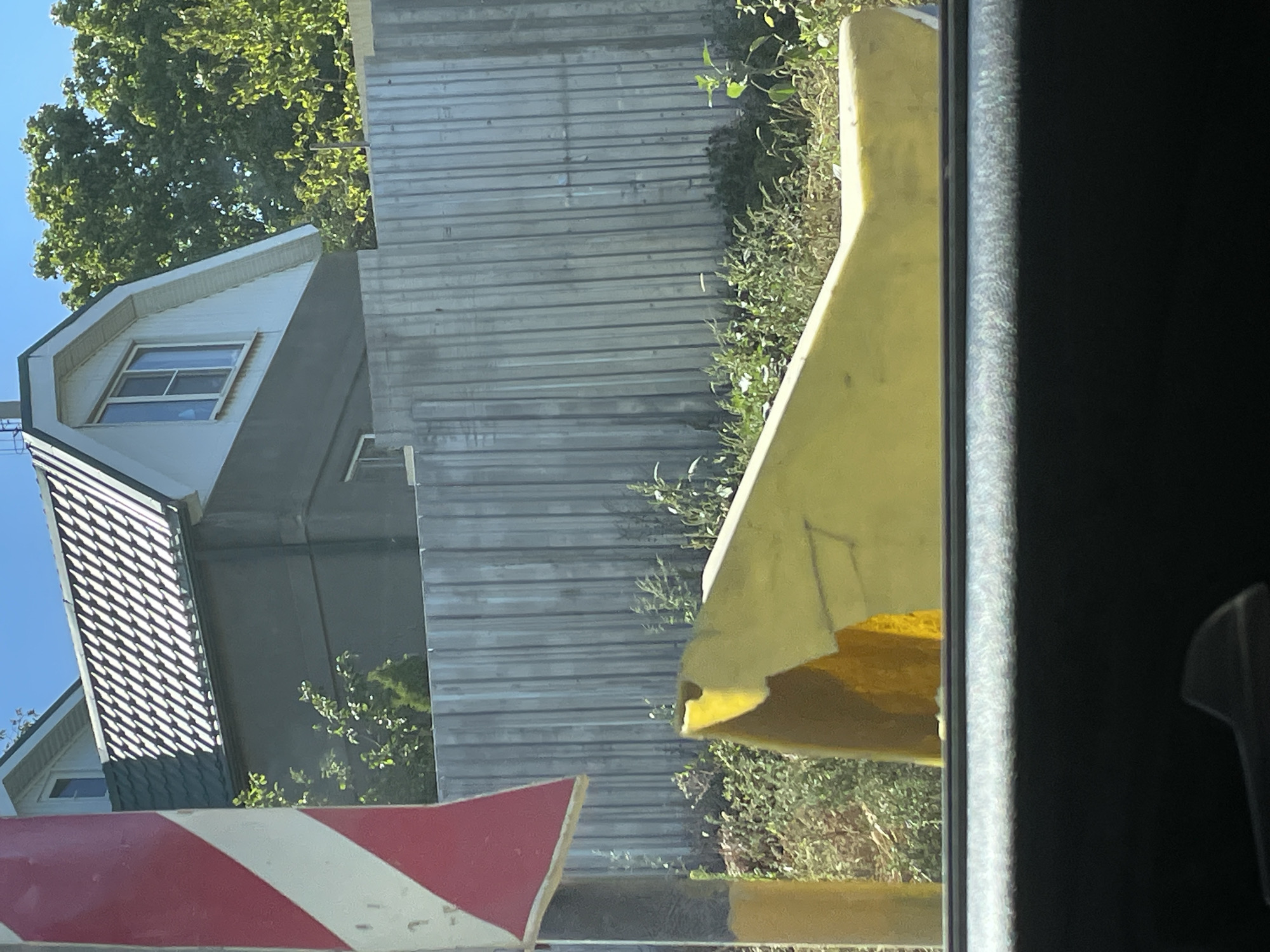 The image depicts a view from inside a vehicle looking out towards a residential area. In the foreground, the dark interior of the car is visible, with a sliver of the passenger seat's texture and a dark handle below the window line. Through the window, a bright yellow, curved object is prominent, appearing to be a piece of discarded plastic or foam. Beyond this, a weathered, corrugated metal fence runs horizontally across the frame, partially obscuring the background. Behind the fence, a two-story house with a dark grey lower level and a white upper gable section is visible. The house has a dark-tiled roof and a single window on the upper level. Lush green trees and foliage surround the house, suggesting a suburban or semi-rural setting. To the left of the house and fence, a red and white striped warning sign is partially visible, mounted on a post that also has yellow and grey sections. The sky above is a clear, bright blue. The overall scene suggests an outdoor environment, possibly in a residential area of Rostov-on-Don, Russia, given the typical fencing and architectural style observed.