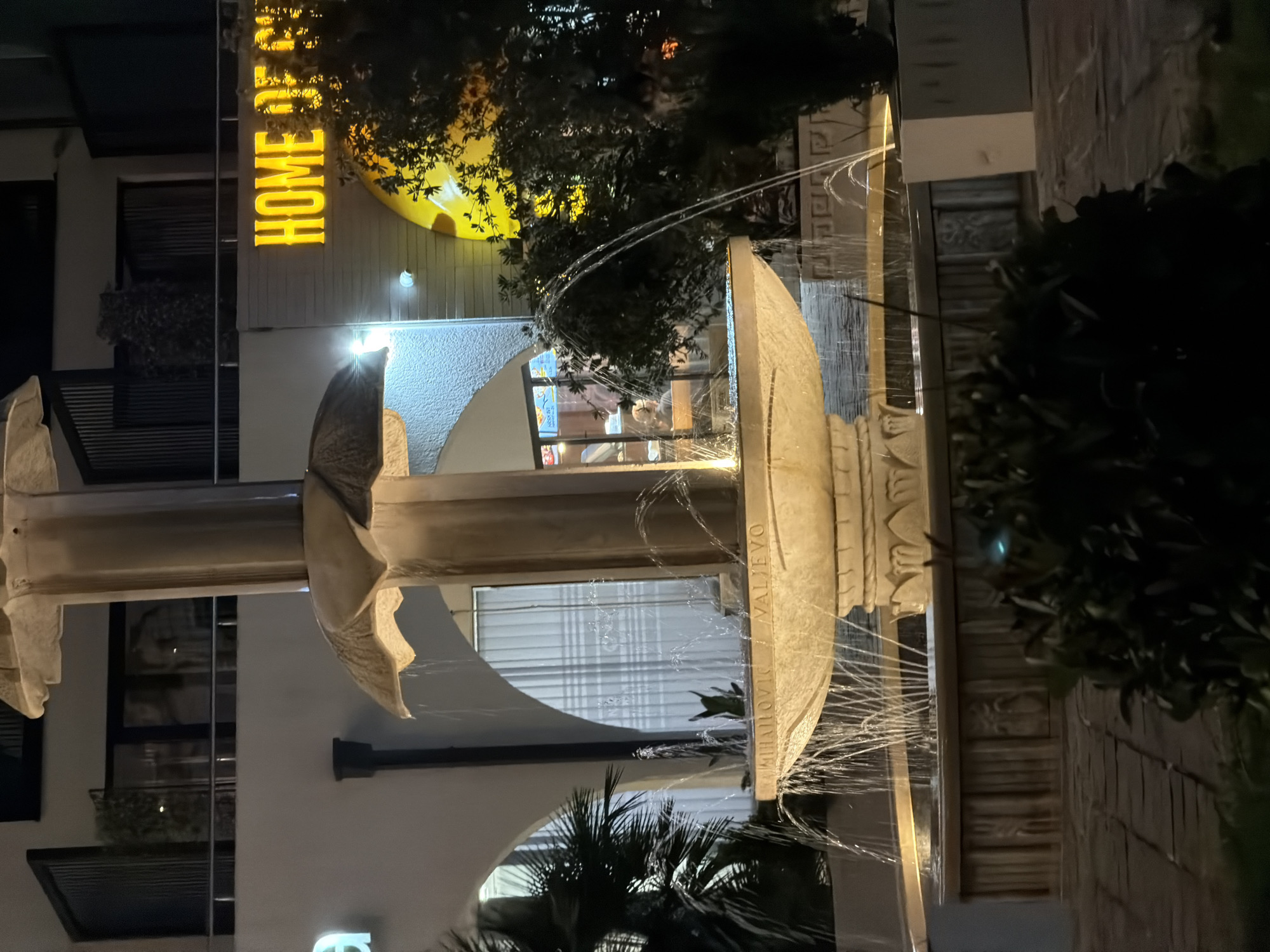 An illuminated fountain is the central focus of this nighttime outdoor scene, situated in front of a building with a prominent yellow sign reading "HOME OF CH." The fountain, made of light-colored stone, features multiple tiers with water cascading down. Text is inscribed on the fountain's basin, partially visible as "MIHAILOVIC VALJEVO."
Behind the fountain, the building is lit, revealing arched windows with sheer curtains. A palm tree is visible to the left, and dense foliage is present to the right, partially obscuring the building. The ground in front of the fountain is paved with stone. The overall ambiance suggests an evening setting, possibly a public space or a commercial establishment. The location is identified as Budva, Montenegro.