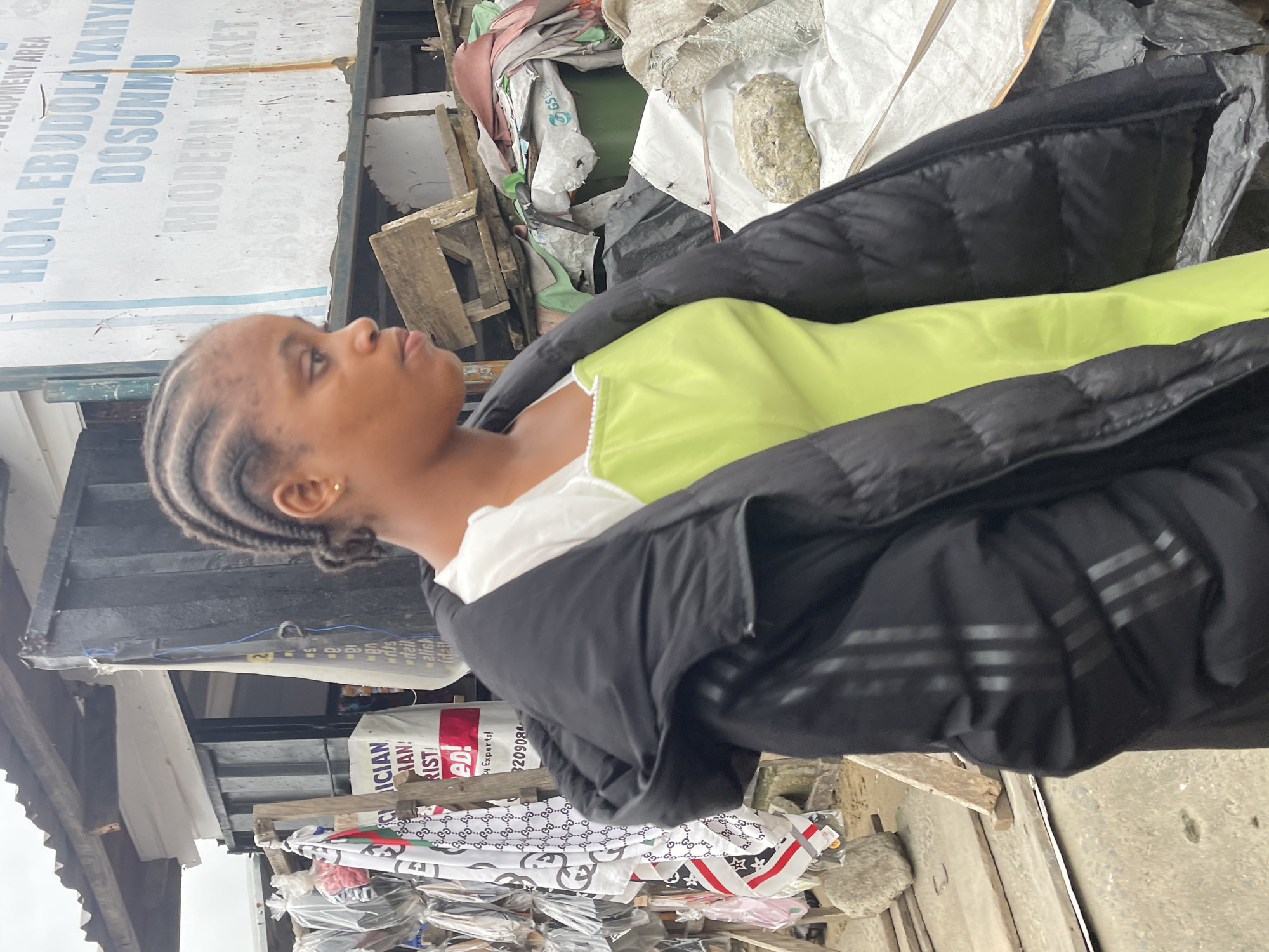 The image depicts a young Black woman standing outdoors in what appears to be a market area in Ado, Nigeria. She is wearing a black puffer jacket over a bright green dress with white trim. Her hair is styled in braids. Behind her, a white sign with blue lettering is visible, with the words "HON. EBUDOLA YAHYA DOSUNMU" and "MODERN MARKET" being legible. To her left, an assortment of items, possibly fabrics or clothing, are displayed on racks. The background suggests an informal market setting with structures, debris, and what looks like tarps or plastic sheeting. The overall scene is somewhat cluttered and appears to be taken during daylight hours.