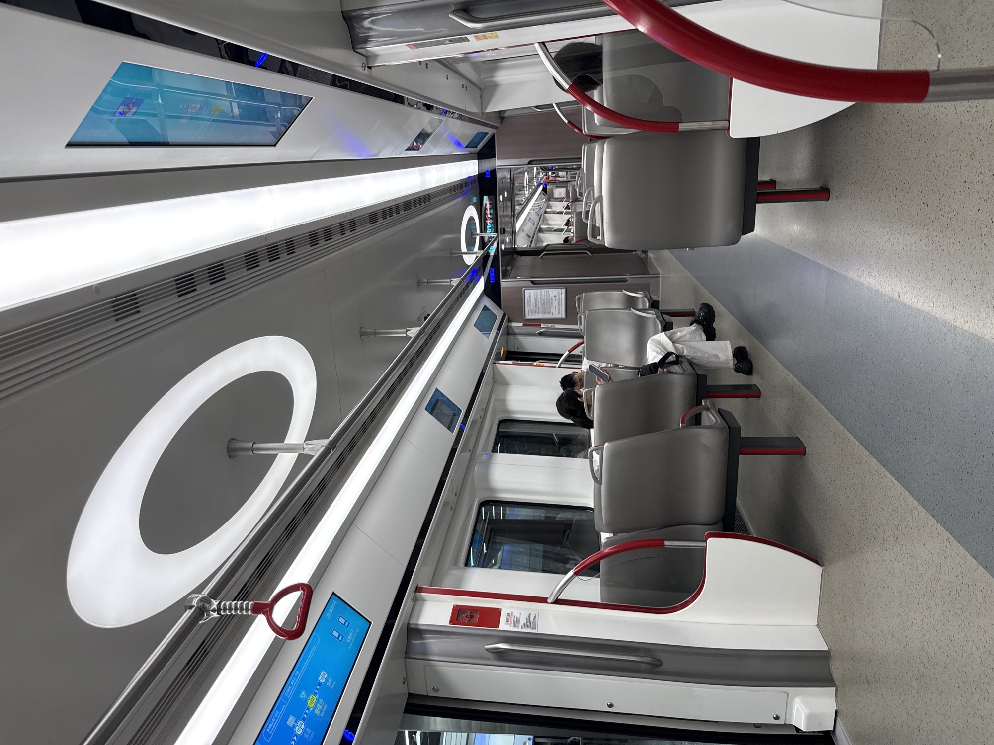 The image depicts the interior of a modern subway car. Seating consists of gray-colored chairs with red handrails. The walls are white and gray, featuring large circular light fixtures and digital screens displaying transit information. There are windows running along the sides of the car. A few people are visible sitting in the chairs. The subway car appears clean and well-maintained. There is a station information poster on the subway. Based on the information on the screen, the location is most likely Haizhu District, China.
