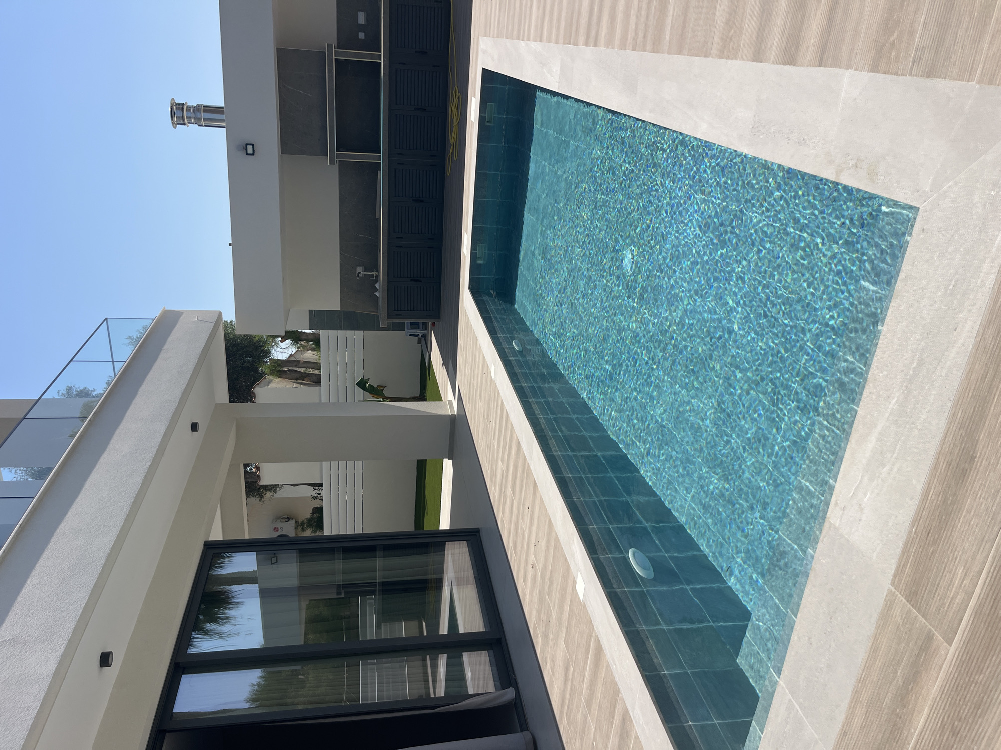 The image depicts a modern outdoor swimming pool area adjacent to a residential building. The pool itself is rectangular with turquoise water, tiled interior, and a light-colored coping. The surrounding deck is paved with light-brown wooden planks and the same light-colored stone used for the pool coping. To the left, a large glass sliding door opens from the building, reflecting palm trees and the sky. The building has a white facade with a balcony on the upper level, featuring a glass railing. To the right, an outdoor kitchen area is visible, with dark cabinets and a gray tiled backsplash, topped by a stainless-steel chimney. The environment is sunny with a clear blue sky. The location is the Moni Community, Cyprus. There are no people or animals visible in the image.