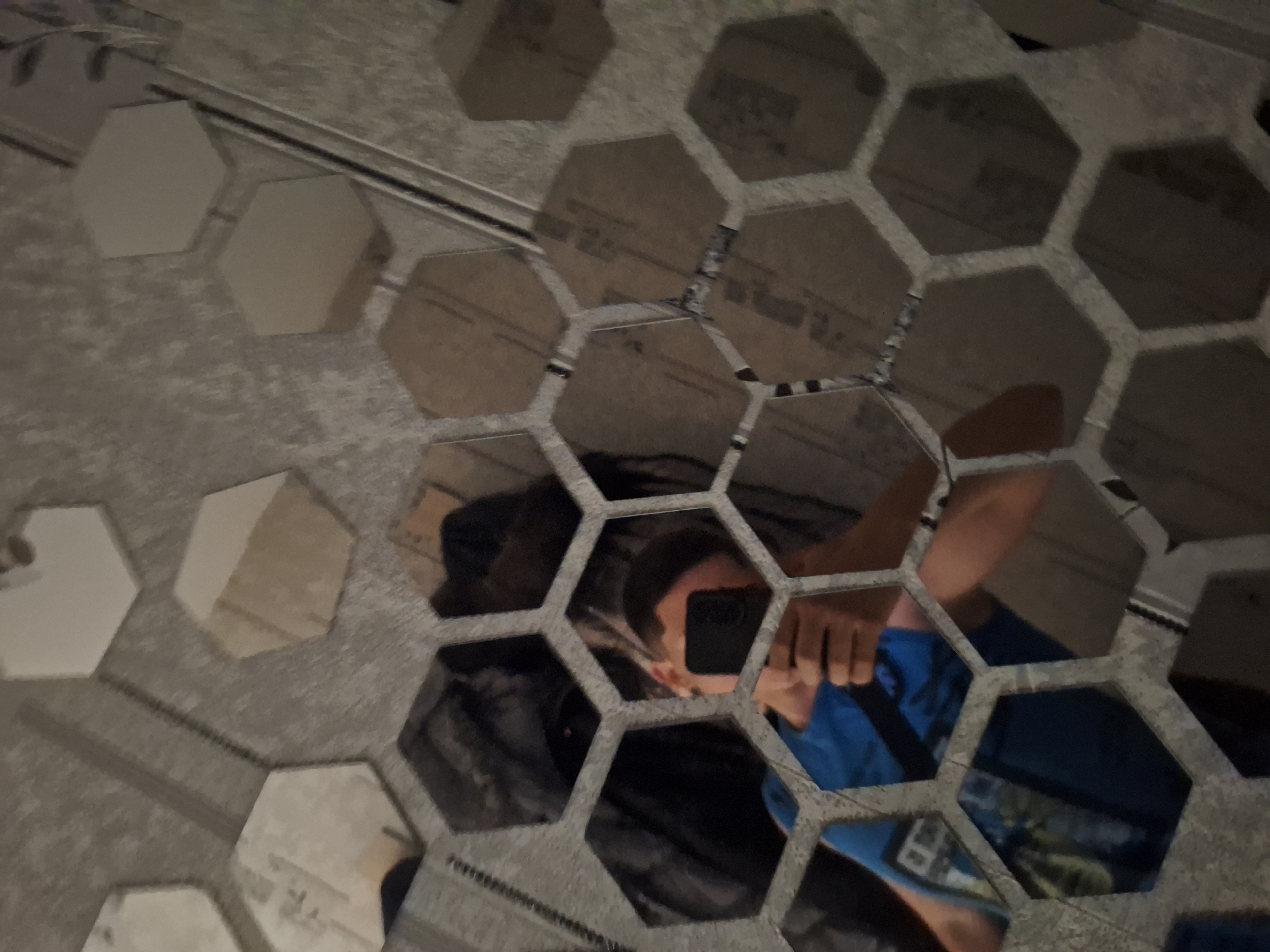 The image displays a close-up of a wall decorated with hexagonal mirrors. The mirrors are arranged in a honeycomb pattern, reflecting various objects and a person. The person, a young individual, is visible in the center of the image, holding a smartphone to take a selfie. They are wearing a blue t-shirt. The background reflection shows what appears to be clothing and possibly furniture. The wall itself has a textured, possibly wallpapered, finish. The overall lighting is dim, contributing to the dark tones of the reflections. There is no discernible information within the image that would indicate its location in Varna, Bulgaria.
