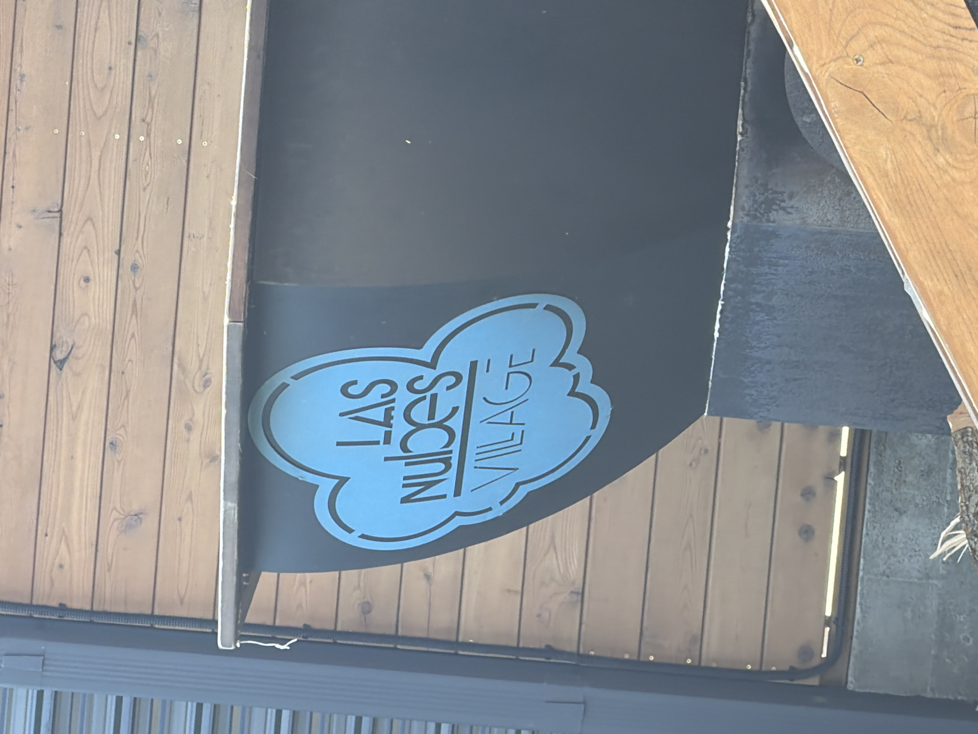 The image shows a close-up of a sign with a blue cloud outline and text in Russian. The text reads "LAS NUBES VILLAGE". The sign is mounted on a dark surface, possibly metal or painted wood, which is attached to a wooden wall. The wooden wall is made of horizontal planks. To the left of the sign is a vertical dark grey ribbed pipe or conduit. Below the sign is a concrete structure. In the lower right corner, a piece of angled wooden planking is visible. The scene appears to be outdoors. The sign is the primary subject of the image.