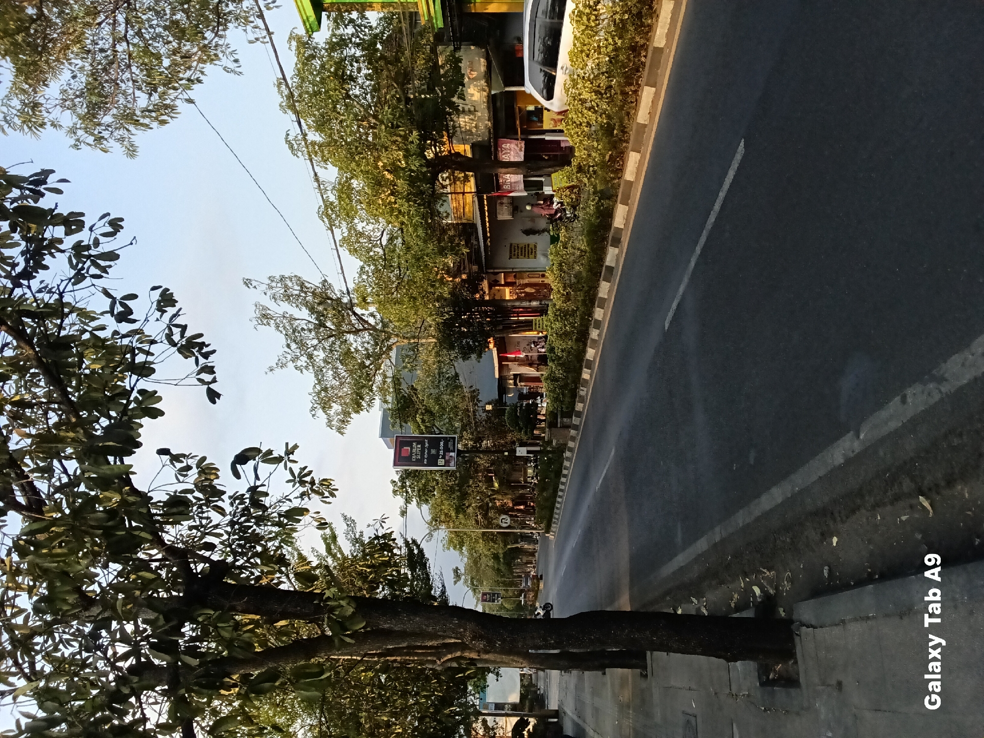 The image shows a street scene in Kelutan, Indonesia. The street is lined with trees on the left sidewalk and a row of buildings and more trees on the right. The road is paved and has lane markings. Several vehicles are visible, including a white car parked on the right and a motorcycle on the left side of the road in the distance. There are also people on the sidewalk and in front of some of the buildings. Signs are posted along the street, including one for "DIARUM SUPER" and another with Indonesian text. The sky is clear and blue. The overall impression is of a typical urban street in a tropical or subtropical climate.
