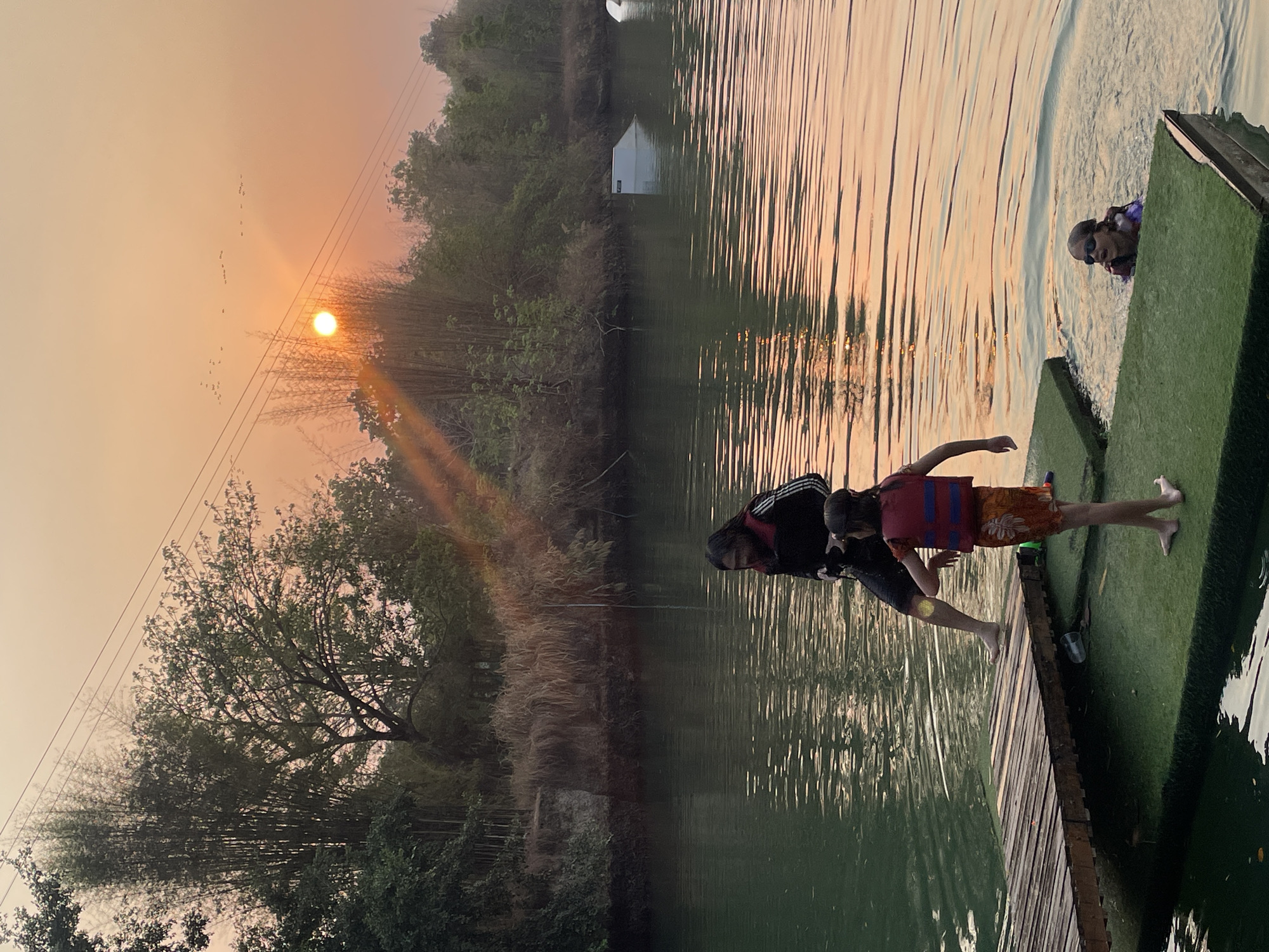 The image depicts a lakeside scene at dusk in Tambon Nam Phrae, Thailand. An orange sun hangs low in the sky, silhouetting a flock of birds and a tree with leafless branches. A wooden pier extends into the calm, reflective water. Two people stand at the end of the pier, one seemingly assisting the other to enter the water. In the water, a third person wearing goggles and a flotation device appears to be swimming. A small, white structure sits further back on the opposite shore, partially obscured by trees.