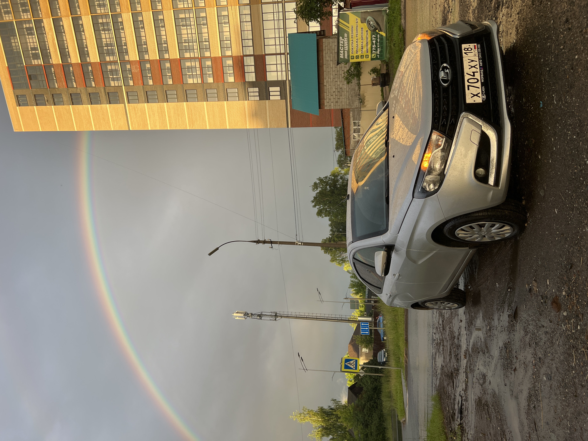 A silver Lada car is parked on a wet, muddy street in front of a multi-story building in Izhevsk, Russia. A rainbow arcs across a cloudy, grey sky. The building is modern, with a yellow brick facade and numerous windows. A cell tower is visible in the background, along with streetlights and trees. A sign for "Master Shinn" (Master Tire) is mounted on a small structure to the right of the car. The car's license plate reads "X 704 XY 18". The scene suggests it has recently rained.