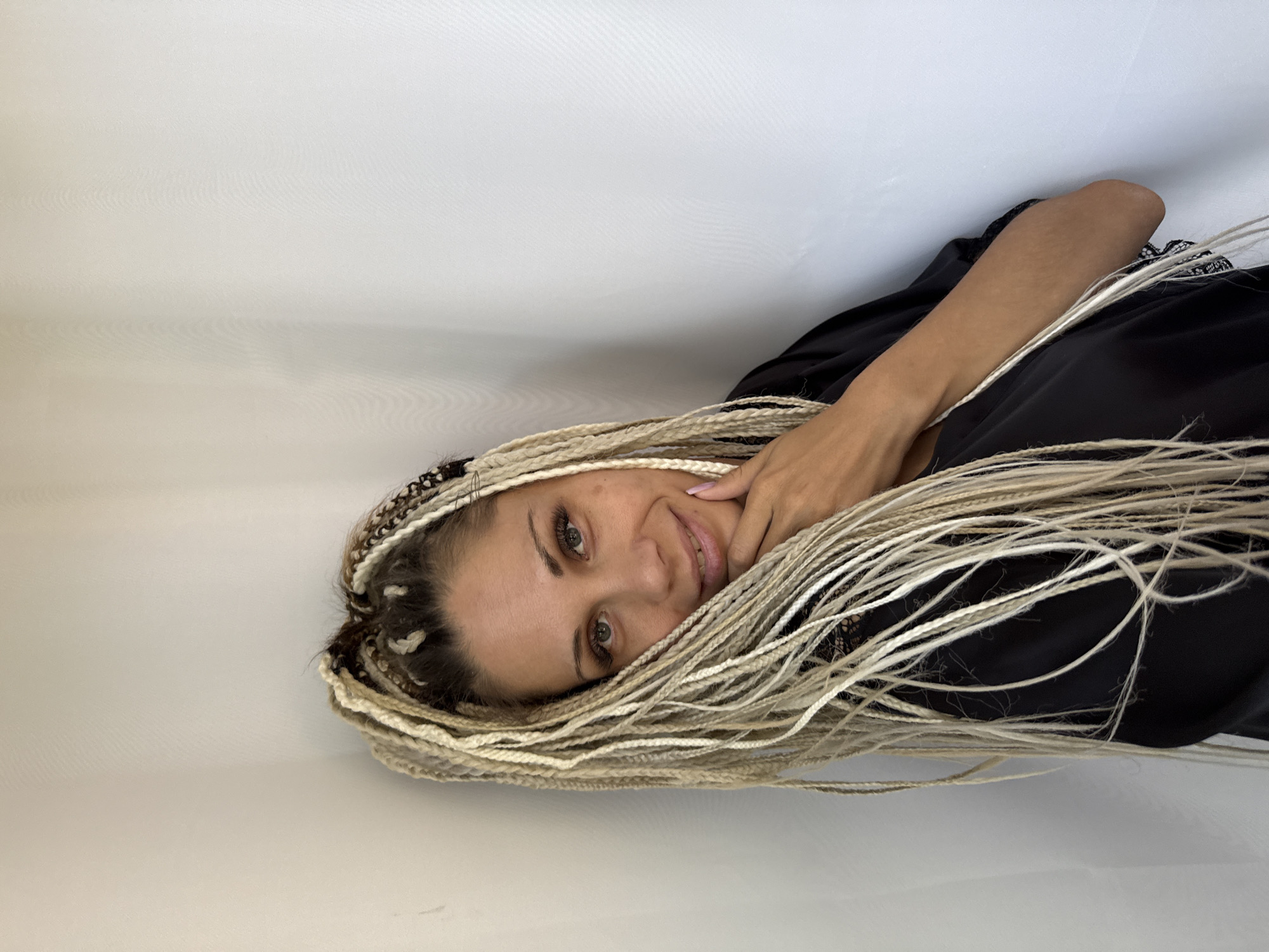 A woman with blonde box braids is leaning against a white wall in what appears to be an indoor setting. She is looking at the camera with a slight smile and has her hand resting on her chin. She is wearing a black garment with lace detailing. The background is a plain white wall. There is no indication of the specific location within Rusanovo, Russia.
