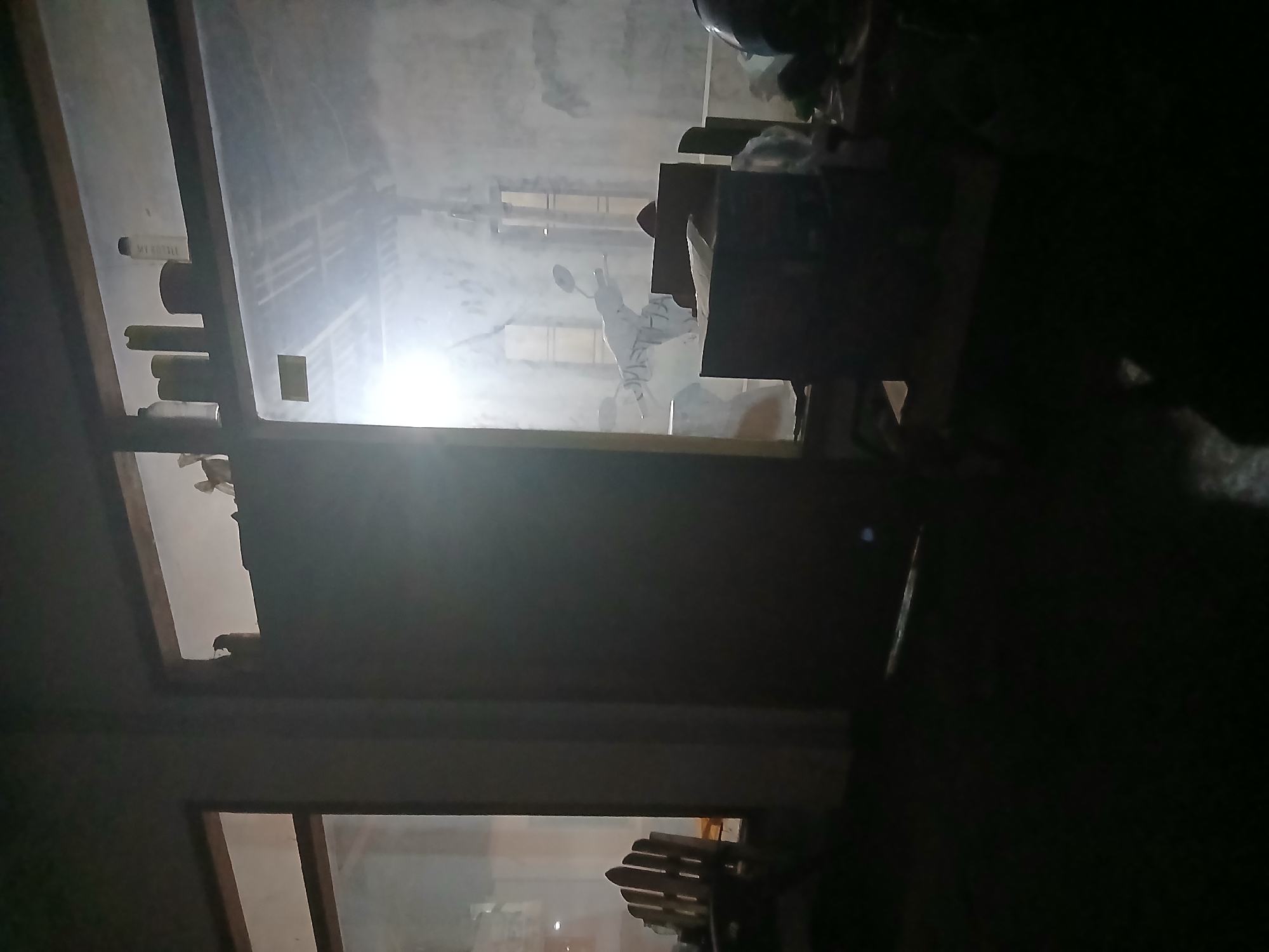 This is a dark, interior shot of what appears to be a garage or storage area in Kademangan Kulon, Indonesia. The image is dominated by shadow, with light emanating from a bright opening, likely a doorway or large window. In the center of the frame, a white moped is partially visible, covered in what appears to be dust or debris. To the right of the moped, several cardboard boxes are stacked, and to the far right, a dark helmet is visible. On a shelf above the moped, several cylindrical objects, possibly bottles or cans, are arranged. To the left of the main area, a wooden Adirondack chair is positioned near a window. The overall scene is cluttered and dimly lit, suggesting a neglected or disused space.