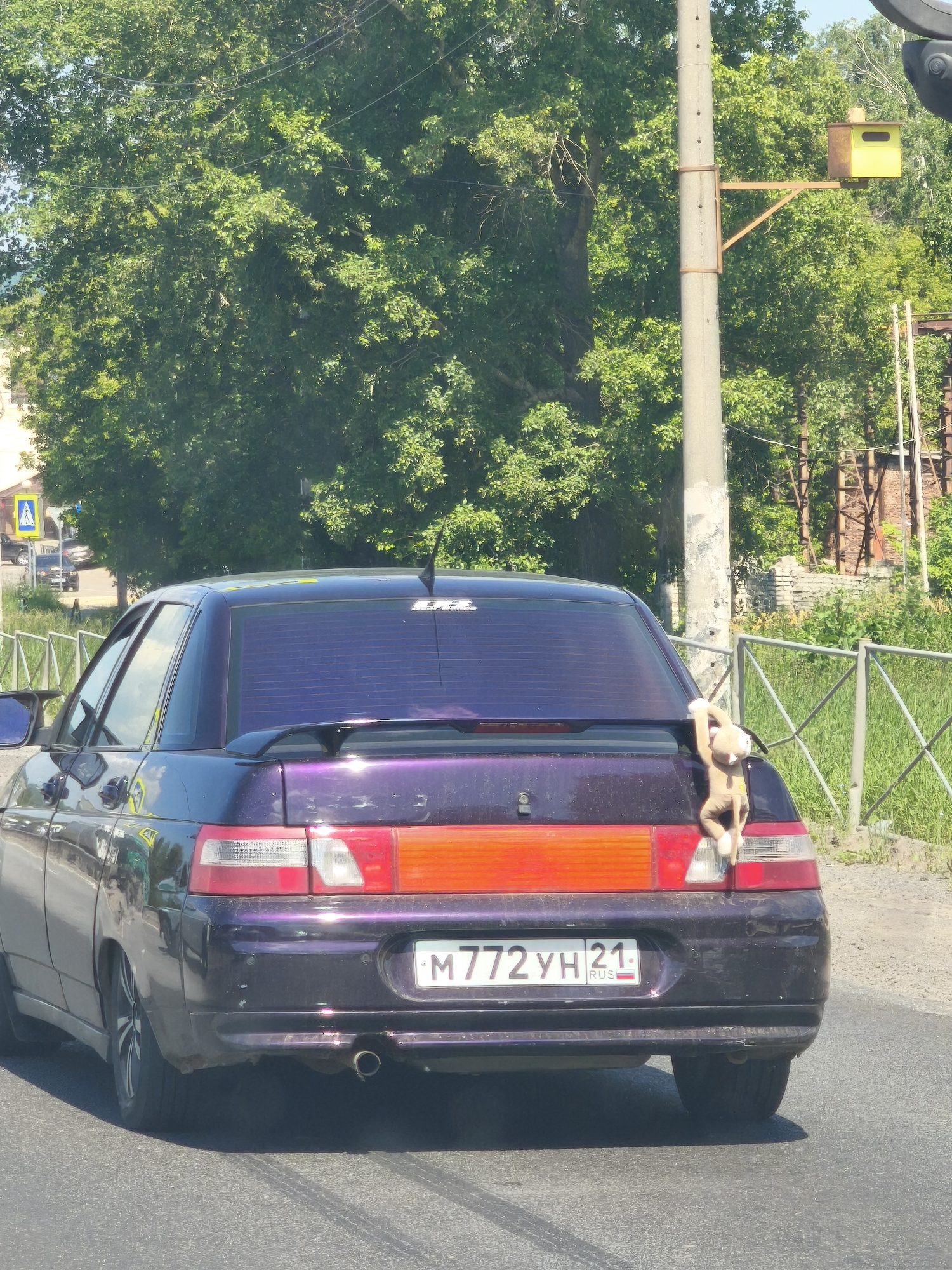 A purple sedan is parked on a paved road in Tsivilsk, Russia. The car has a spoiler on the trunk and a stuffed monkey hanging from its rear. The license plate reads "M772YH 21 RUS". The background features trees, a fence, and a utility pole with a yellow traffic camera mounted on it. The sky is clear and blue.
