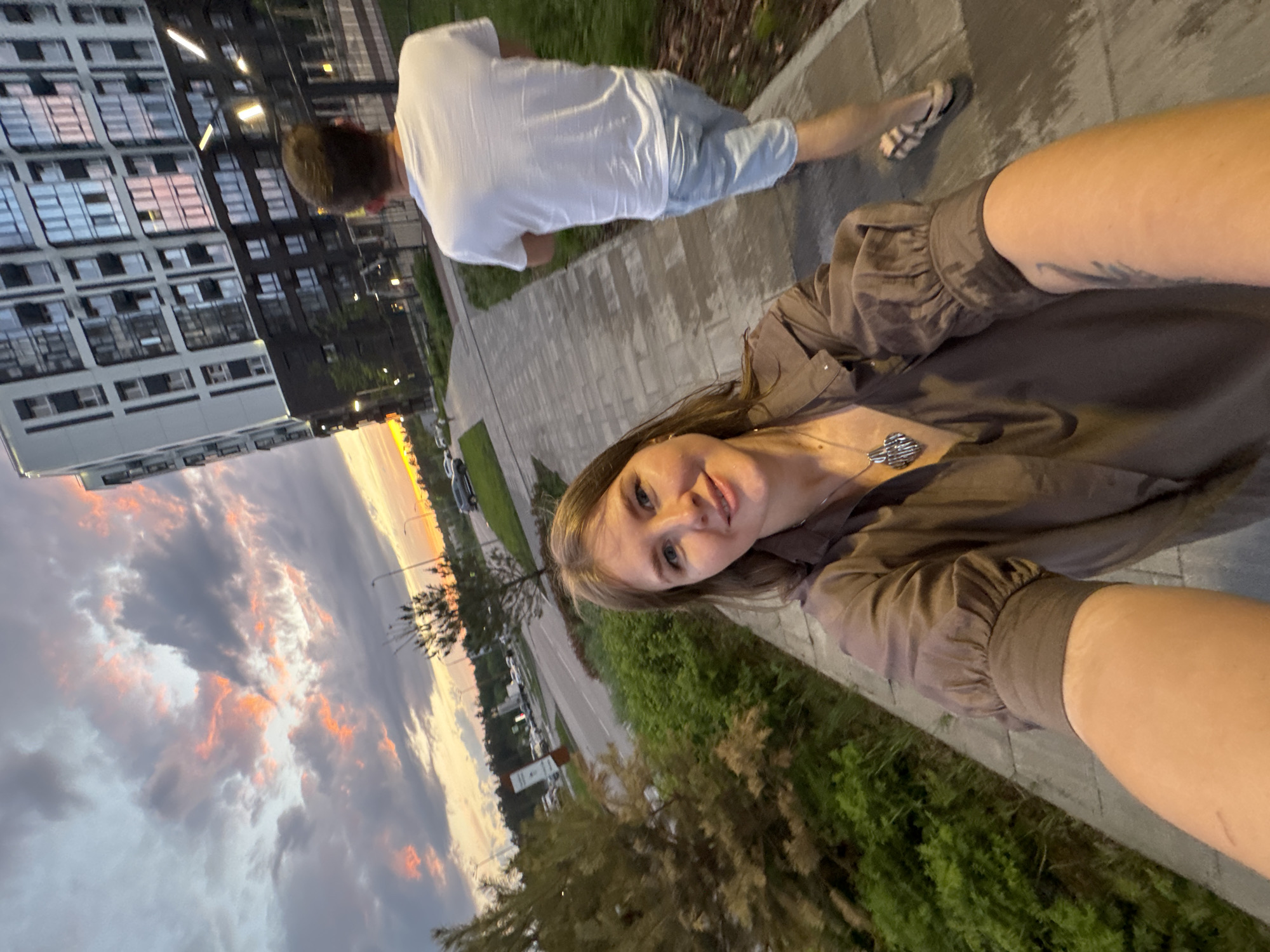 A selfie of a woman in Perm, Russia on a sidewalk with a building in the background and a sunset visible. A man wearing a white t-shirt and jeans is walking ahead of her. The woman is wearing a brown blouse and a necklace. There are trees, a street, and parked cars visible in the background.