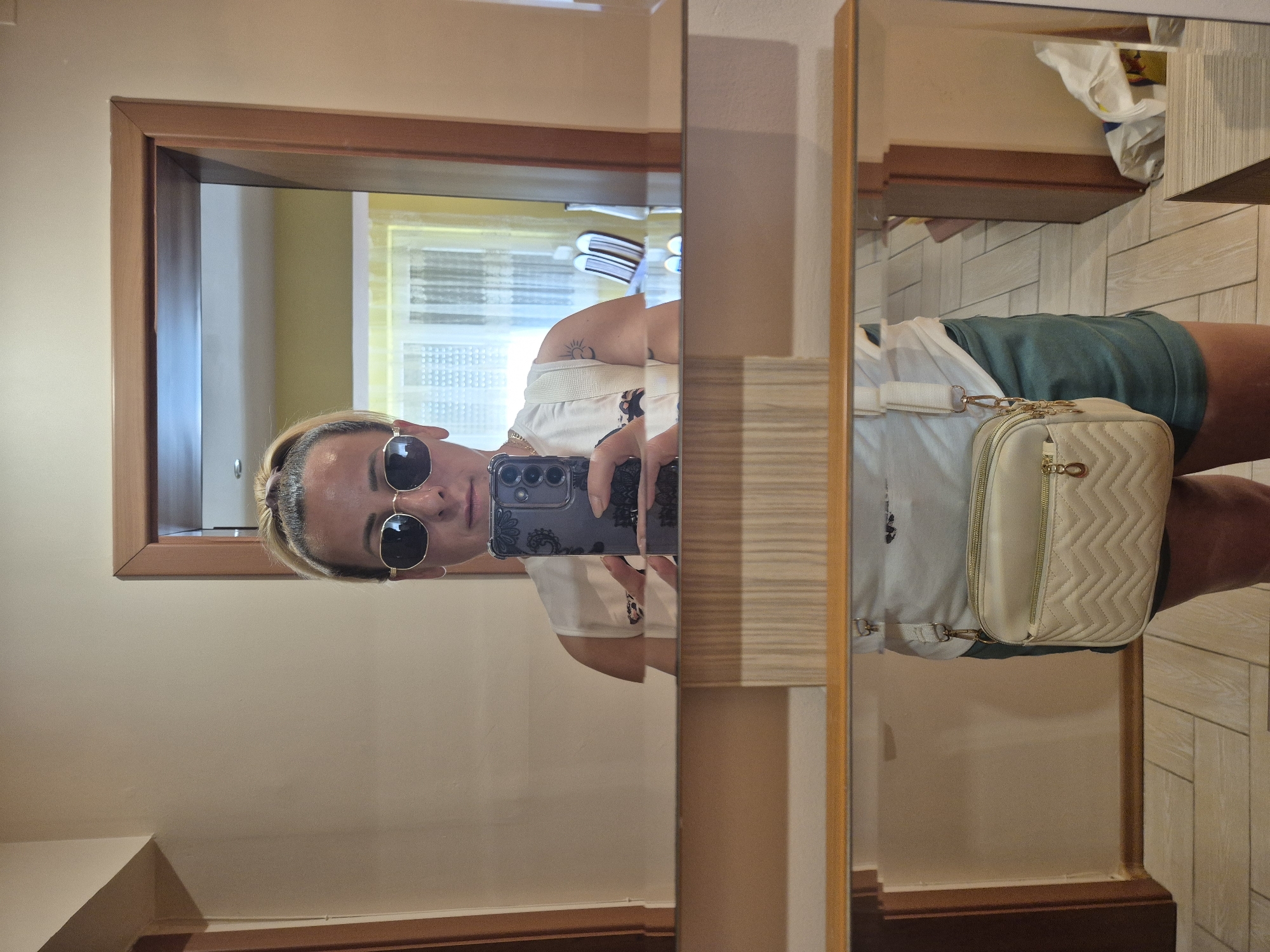 A person is taking a mirror selfie in a hallway. They are wearing sunglasses, a white sleeveless top, and dark green shorts. A cream-colored crossbody bag is hanging from their shoulder. The hallway has beige walls and light-colored tiled flooring. A doorway with a wooden frame is visible on the right side of the image, and a mirror reflects the room behind the person. The room in the reflection appears to be a living space with furniture and curtains.