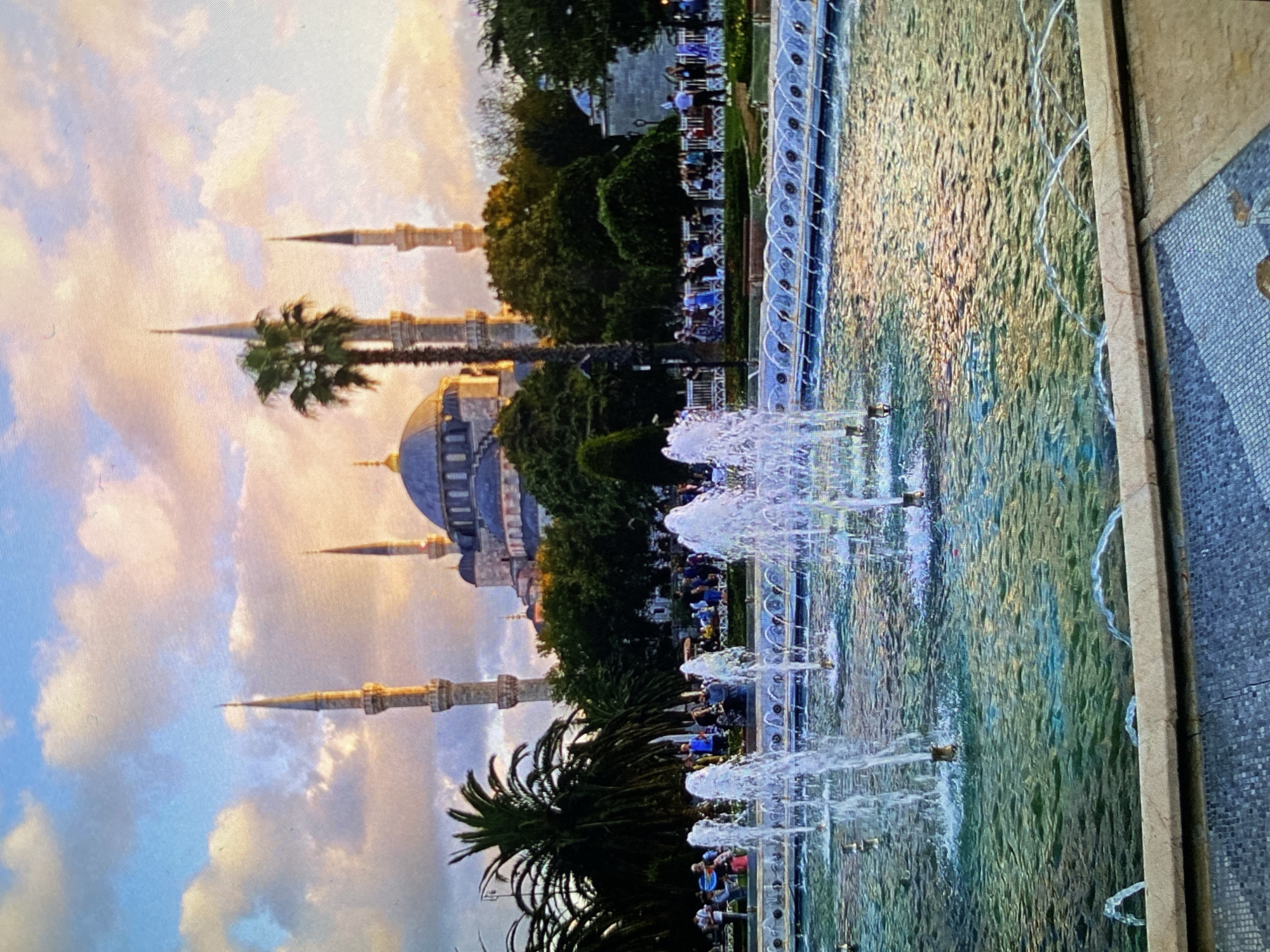 The image, initially rotated 90 degrees counter-clockwise, depicts a grand mosque with multiple minarets and a prominent dome, strongly resembling the Sultan Ahmed Mosque (Blue Mosque) in Istanbul, Turkey. This visual evidence contradicts the provided location context of Bankya, Bulgaria.
The scene captures a beautiful moment, likely during the golden hour of either sunrise or sunset, with a soft, warm light illuminating the sky. The sky features scattered clouds painted with hues of pink, orange, and light blue. In the background, the mosque's distinctive grey-blue dome and several slender minarets rise majestically.
In the midground and foreground, a long, rectangular fountain stretches across the frame, actively spraying water upwards from multiple jets, creating shimmering white plumes. The water in the fountain reflects the warm light, appearing in various shades of green, blue, and gold. Lush green trees, including several tall palm trees, are interspersed around the mosque and along the sides of the fountain, framing the architectural marvel.
Numerous people are visible throughout the public space, casually dressed, either strolling or gathered in groups around the fountain and among the trees. Some appear to be seated at outdoor tables. The overall atmosphere is peaceful and serene, suggesting people are enjoying a pleasant evening in this historic and scenic location. No discernible text is visible in the image.