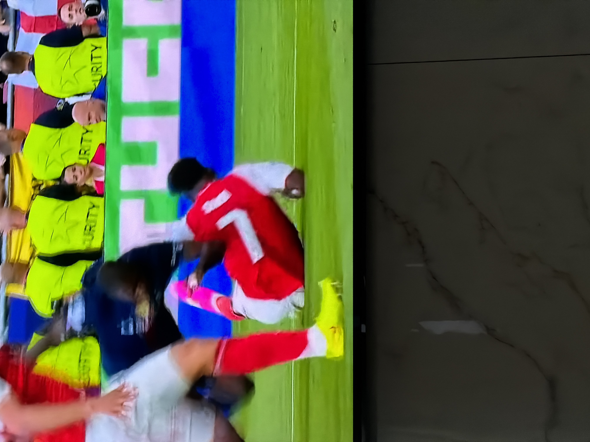 A television screen displays a soccer game in progress, with players visible in red and blue uniforms. Security personnel in yellow vests line the perimeter of the field. The location is not discernible from the image.