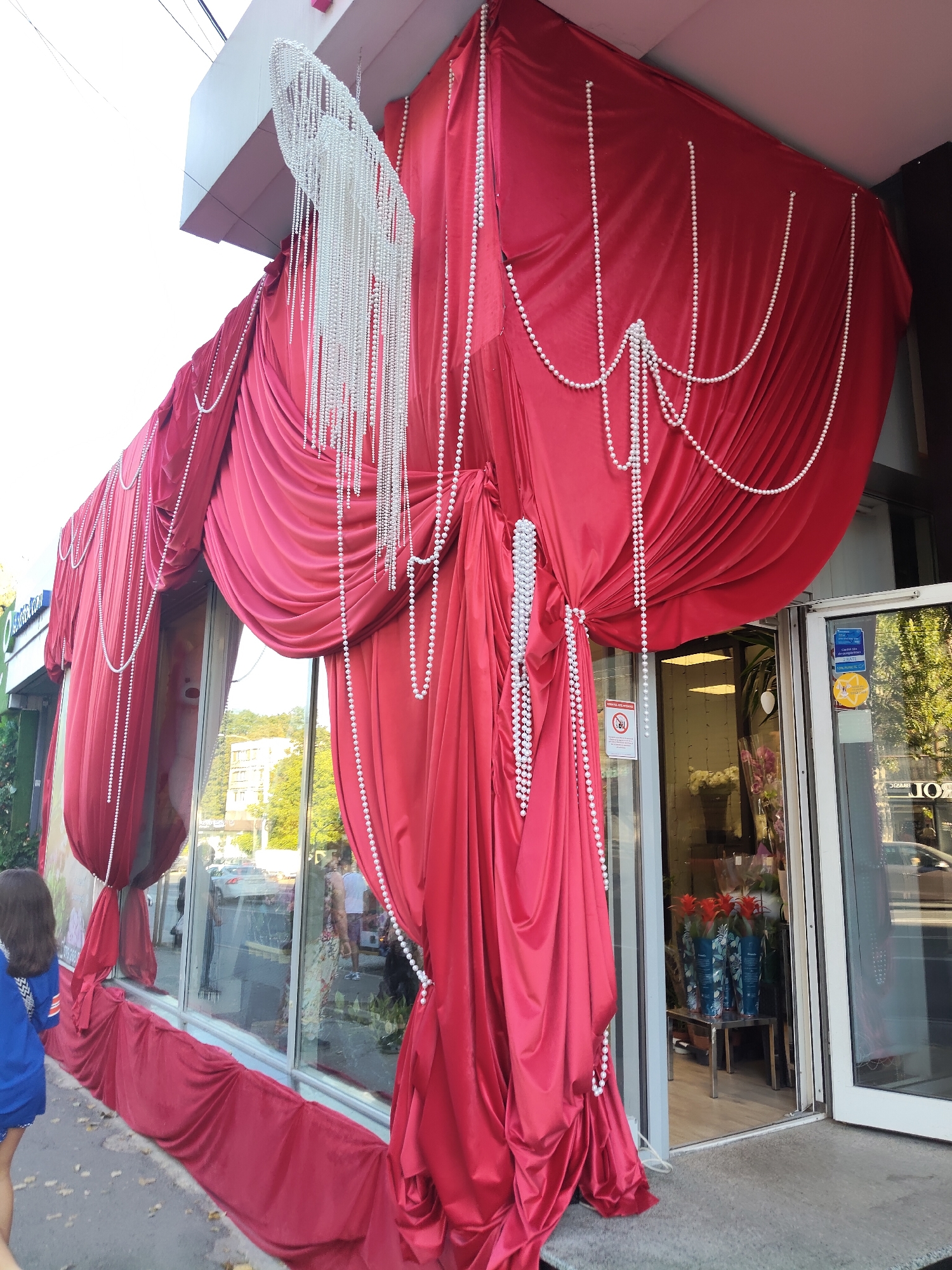 This image depicts the facade of a shop in Chișinău, Moldova, decorated with opulent red drapery and strings of faux pearls. The shop's entrance and large display windows are partially obscured by the elaborate fabric. The red drapes are arranged in dramatic folds, cascading from a structure above the shop and extending down to the sidewalk. Pearl strands are intertwined with the fabric, creating a decorative effect. Through the windows, a glimpse of the interior is visible, showing floral arrangements and other merchandise. The exterior environment includes a sidewalk, street, and buildings across the street, suggesting an urban setting. A few individuals are present, including a child in the foreground and people inside the shop. The overall impression is one of a decorated storefront, possibly for a special occasion or as part of a business's visual merchandising.