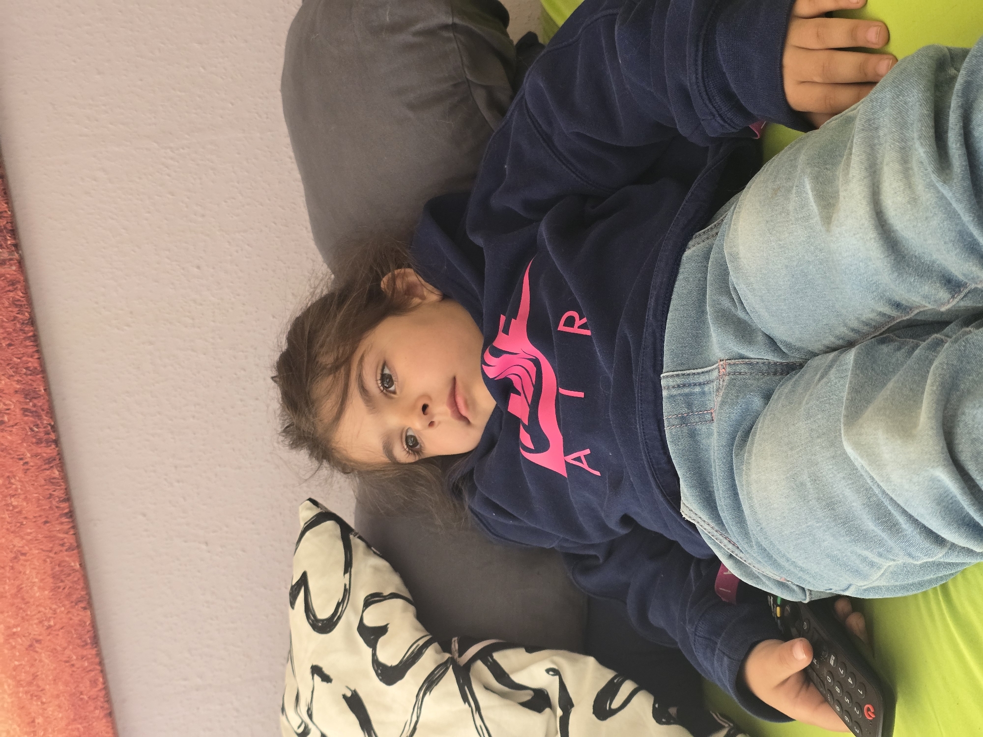 A young girl is lying on a bed, possibly in a residential setting. She is wearing a dark blue sweatshirt with a pink design and light blue jeans. She is holding a black remote control in her right hand. Her head is resting on a pillow, and there is another pillow behind her. The bed has a green sheet, and the wall is light-colored. The image does not provide any information to confirm or deny that the location is Zirndorf, Germany. The scene is indoor and depicts a child relaxing on a bed.