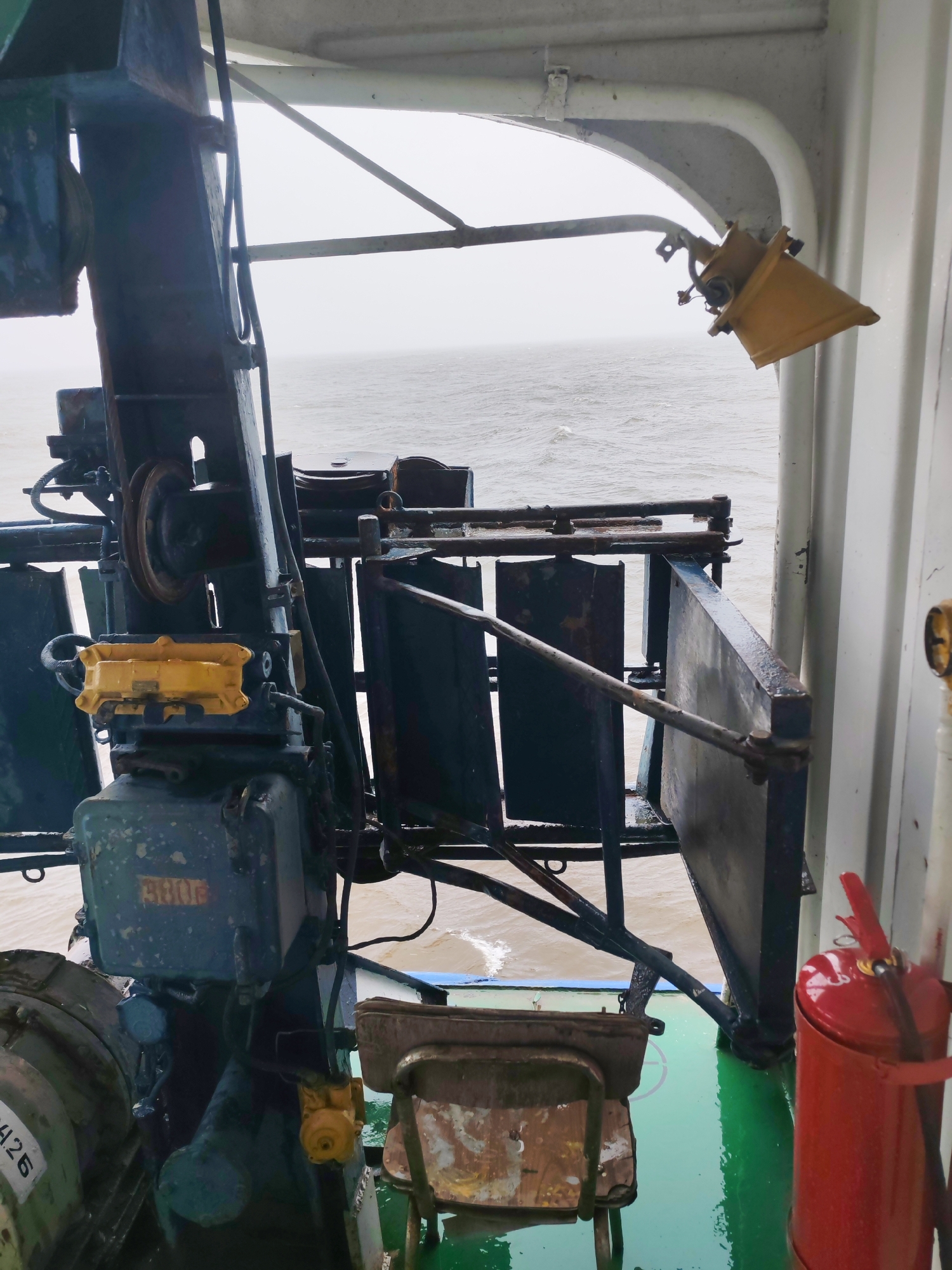The image captures a detailed view of the weathered equipment and structures on a working vessel at sea, likely near Shcheleiki, Russia. A large, dark-colored, robust piece of industrial machinery, possibly a winch or part of a crane, dominates the left and central portions of the frame. This machinery shows significant wear and rust, featuring pulleys, heavy cables, and various metal components, including two distinctive yellow-painted housings – one larger and rectangular on the lower section, and another smaller one higher up.
To the right of the main machinery, a metal barrier or guard rail composed of dark vertical panels provides a boundary before the open sea. The water in the background is choppy under a uniformly gray, overcast sky, suggesting poor visibility and potentially harsh weather conditions. The vessel's deck, visible in the foreground, is painted a bright green and appears wet with standing water, indicating rain or sea spray. A rusty, worn metal chair or stool sits on the deck near the bottom center.
On the far right, a vibrant red fire extinguisher is mounted against a clean, white bulkhead. Above it, a yellow, conical light fixture hangs from an arched overhead structure, which is also painted white or light gray. The clean, contrasting white paint of the ship's interior structures highlights the industrial nature and heavy use of the machinery.
Visible text includes "426" on a white label on the lower-left part of the machinery and "3006" faintly marked on a blue-green housing component. No human figures or direct activity are present, but the scene conveys a sense of a rugged, functional maritime environment.