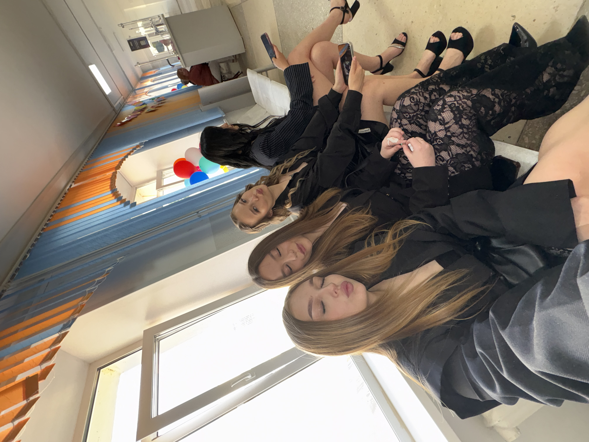 Four young women are seated on a bench in what appears to be a school hallway or common area in Chaikovsky, Russia. The woman in the foreground is looking towards the camera with her eyes closed and lips pursed, as if blowing a kiss, and is wearing a black blazer over a black top and lace pants. To her right, another woman is looking away from the camera with her eyes closed and head tilted. The third woman, from the left, is looking towards the camera with a slight pout and is wearing a black blazer and shorts. The woman furthest to the right is looking to her side and holding a phone. The hallway has a light-colored floor and a white ceiling with fluorescent lighting. Blue and orange vertical blinds are visible along the wall, and balloons are scattered near a window. In the background, a person is seated at a counter. The lighting suggests it is daytime.