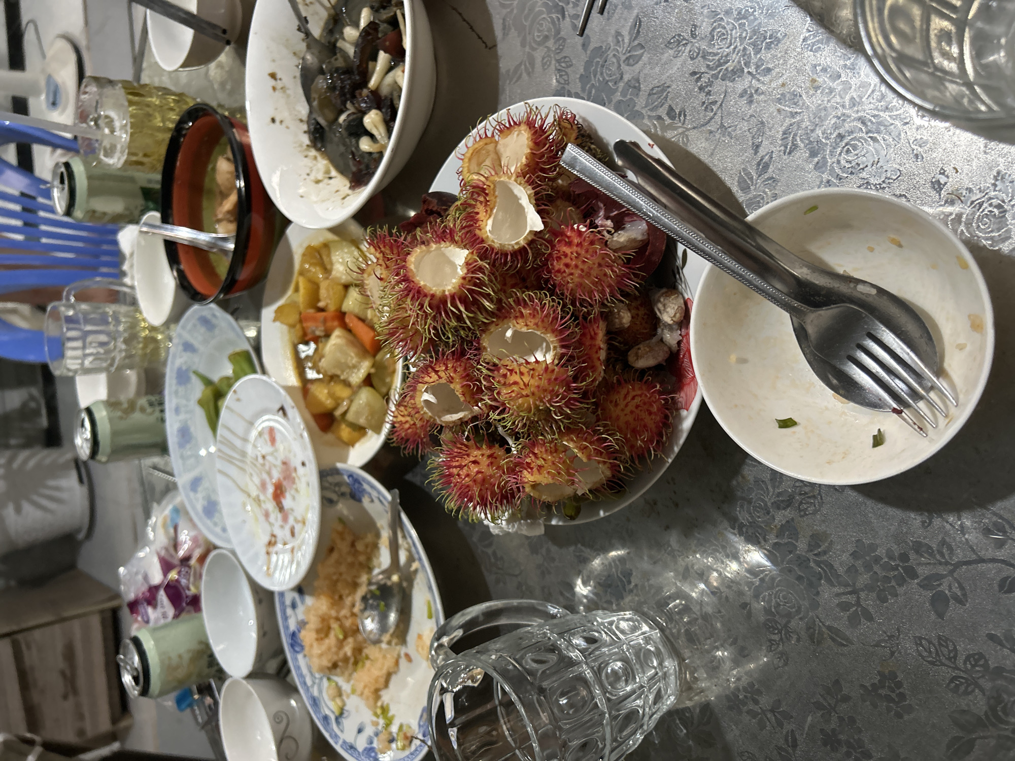 The image depicts a cluttered table at what appears to be a casual meal or gathering, likely in An Hữu, Vietnam. The table is covered with a patterned tablecloth and features a variety of dishes and drinks. In the foreground, a white bowl contains a fork and spoon. To the right of this, a large plate is filled with rambutans, some of which have been opened. Further back, several other plates hold remnants of food, including rice and what appears to be a stew or curry in a black and red bowl. Empty beer cans and glasses are also visible on the table, along with a glass pitcher. The lighting suggests it is evening or indoors with artificial light. The overall scene conveys a sense of post-meal disarray.