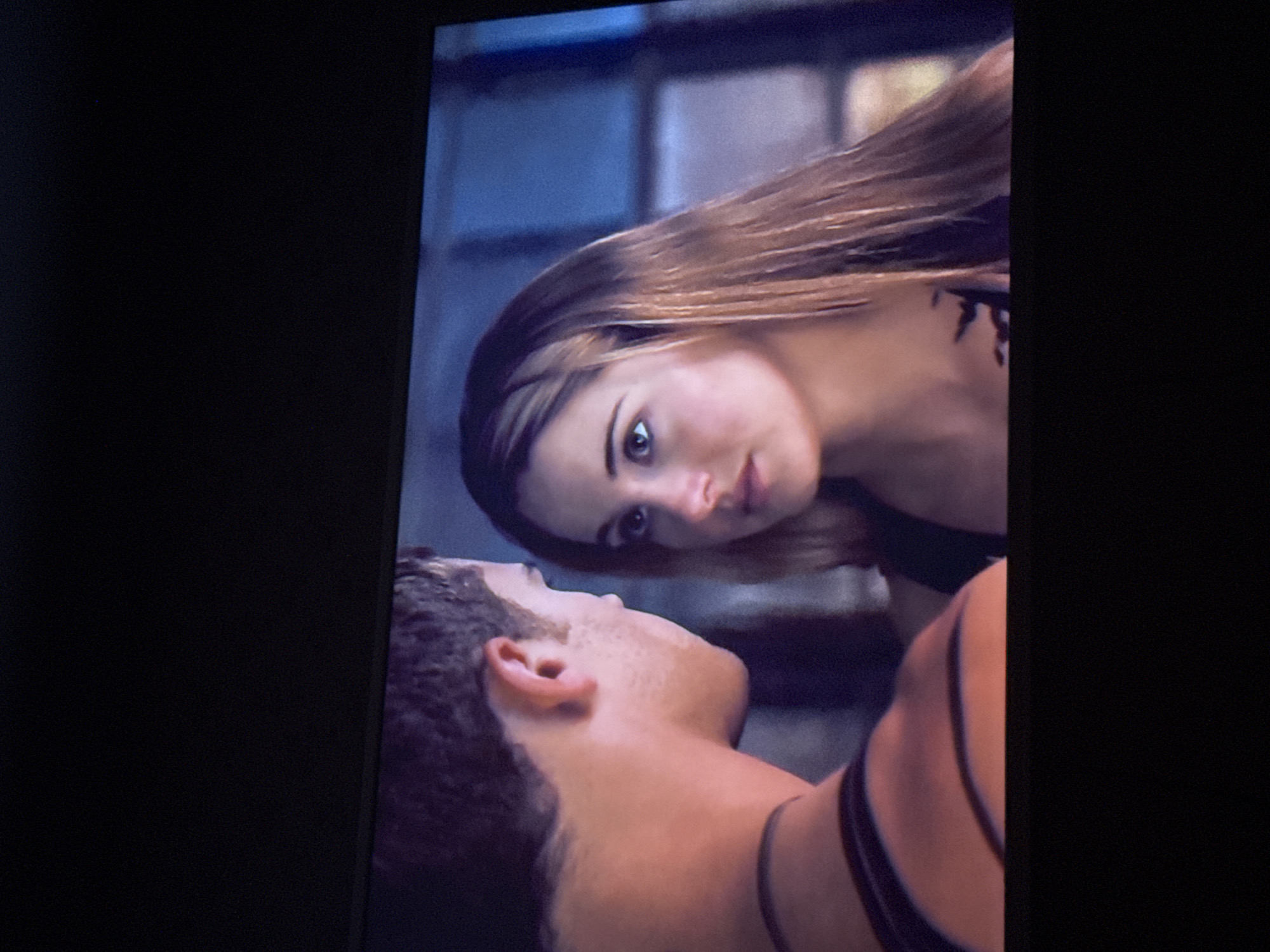 A close-up shot shows a man lying down while a woman leans over him, looking down at him. The woman has light brown hair with darker roots and a fair complexion. A tattoo is visible on her upper chest/shoulder area. The man has short, dark hair and is facing upwards towards the woman. The background appears to be a blurry, indistinct indoor scene with blue-grey tones. There is no specific information to ascertain the location in Mytishchi, Russia.