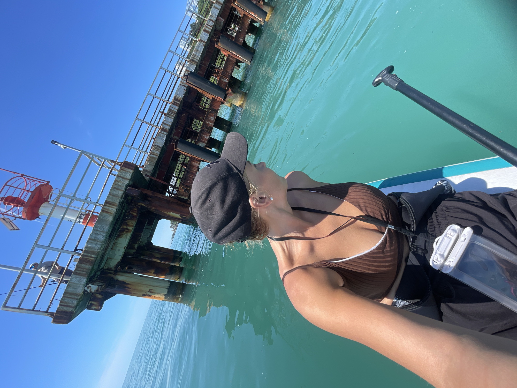 This media file captures a rotated, bright, sunny scene of a person on a paddleboard in clear, turquoise-blue water.
The primary subject is a person, likely female, positioned on a white paddleboard, seen from the chest up. She is wearing a black baseball cap, a brown and white striped halter top, and black shorts. A black waist bag and a white waterproof pouch are visible next to her. A black paddle rests on the paddleboard, extending towards the upper right of the frame.
The activity taking place is paddleboarding near a pier. In the background, an old, robust pier structure, composed of concrete, rusty metal, and wooden piles, extends over the water. Another person can be seen standing on the pier, holding a fishing rod, indicating fishing activity. To the left, a prominent red and white navigational buoy with a metal cage-like structure is visible.
Notable details include the bright blue sky and calm, transparent water, suggesting excellent weather conditions. The time of day is clearly daytime, given the strong illumination. On the red and white buoy, partially visible Cyrillic text "МАРШ" can be discerned. The setting is a coastal area, specifically identified as Sochi, Russia, characterized by the pier and navigational aids. The overall mood conveyed is one of relaxation and enjoyment of the marine environment.