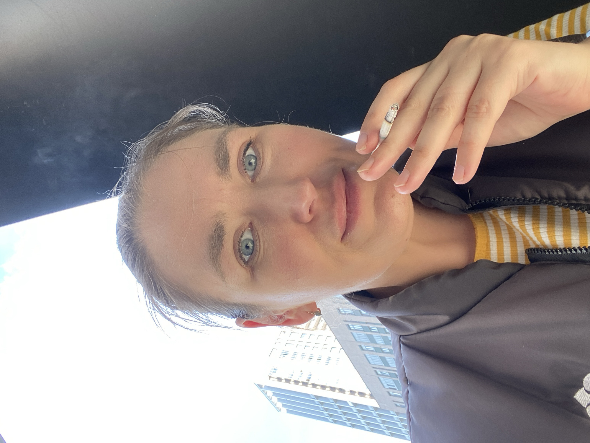 A fair-skinned woman with light blue eyes holds a lit cigarette in her right hand. She is wearing a brown jacket with a yellow-and-white striped shirt underneath. Behind her, a modern high-rise building is visible, partially obscured by the bright sky. The top of the image is black, suggesting an overhead structure, possibly the interior of a car. The location appears to be an urban environment. It's not possible to confirm the location is Yekaterinburg, Russia without more data.