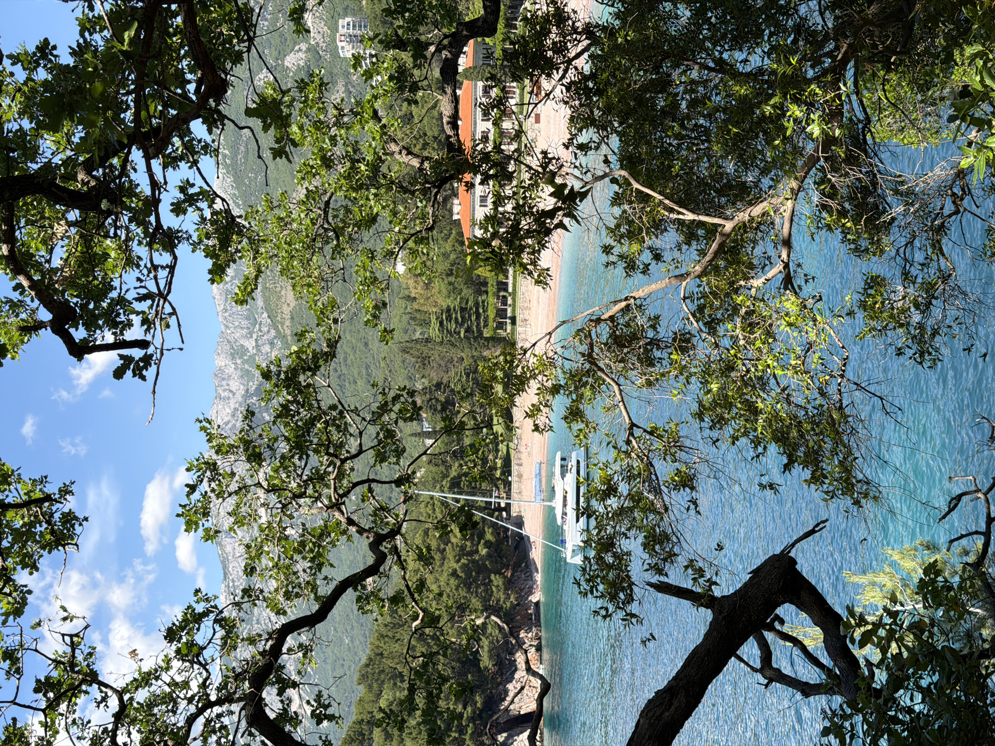 The image depicts a view of the Adriatic coastline in Sveti Stefan, Montenegro, framed by trees in the foreground. In the background, a beach with light-colored sand stretches along the edge of turquoise-blue water. Several people are visible on the beach. Adjacent to the beach are buildings with red-tiled roofs and white walls. Mountains are seen further inland, under a partially cloudy blue sky. A white sailboat is docked near the shore.