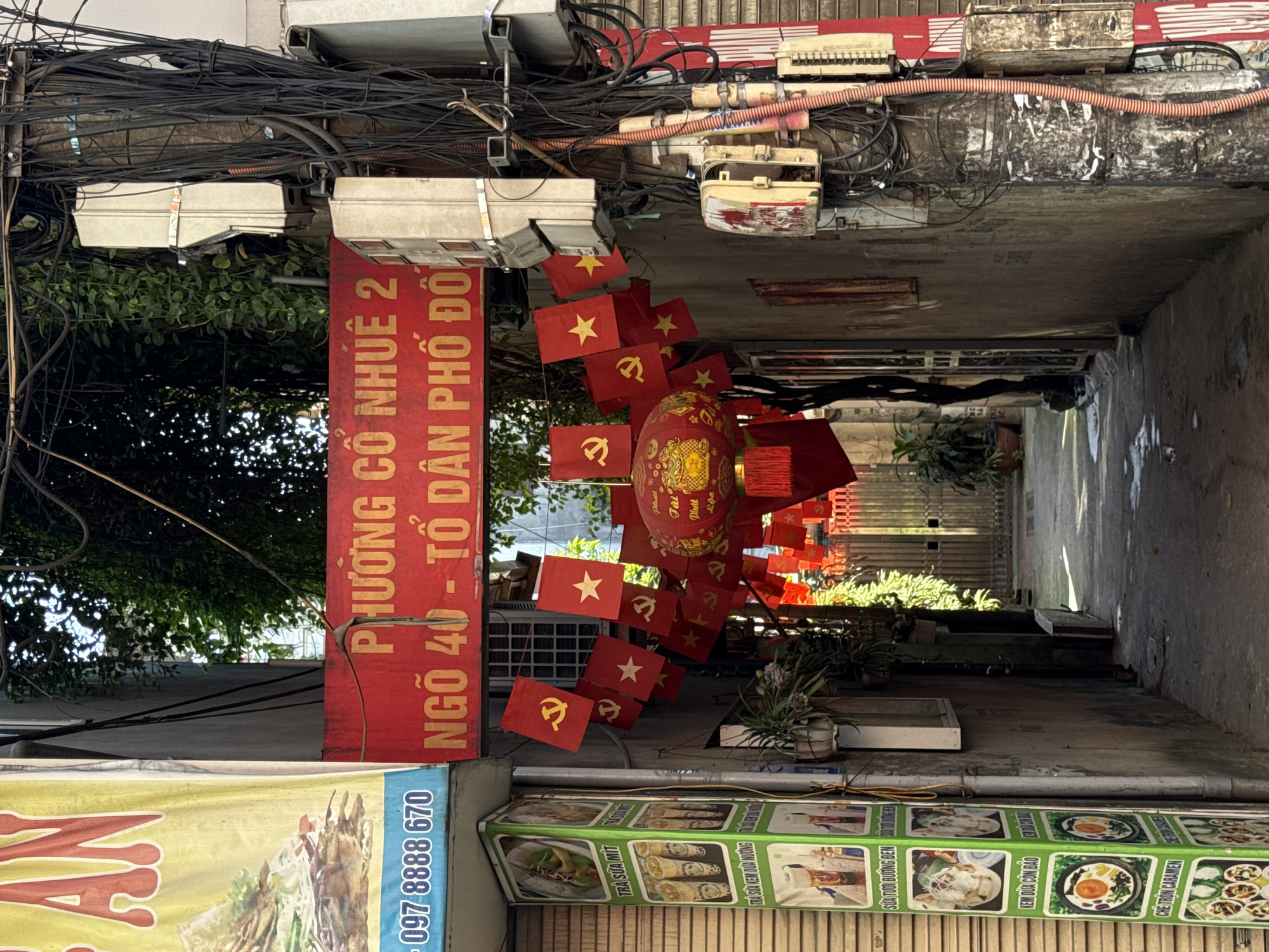 Captured in the city of Hà Nội, Vietnam, the image depicts a narrow alleyway adorned with Vietnamese flags and a traditional lantern. The flags, predominantly red with yellow stars and the hammer and sickle symbol, are strung along the alley, leading towards the depth of the frame. A prominent red banner on the left, written in Vietnamese, provides local orientation. Below, a food advertisement banner in green showcases local cuisine. Overhead, a dense network of wires and utility boxes suggests the urban infrastructure. The alleyway, though narrow, is bright, leading to a garden or courtyard at the end.