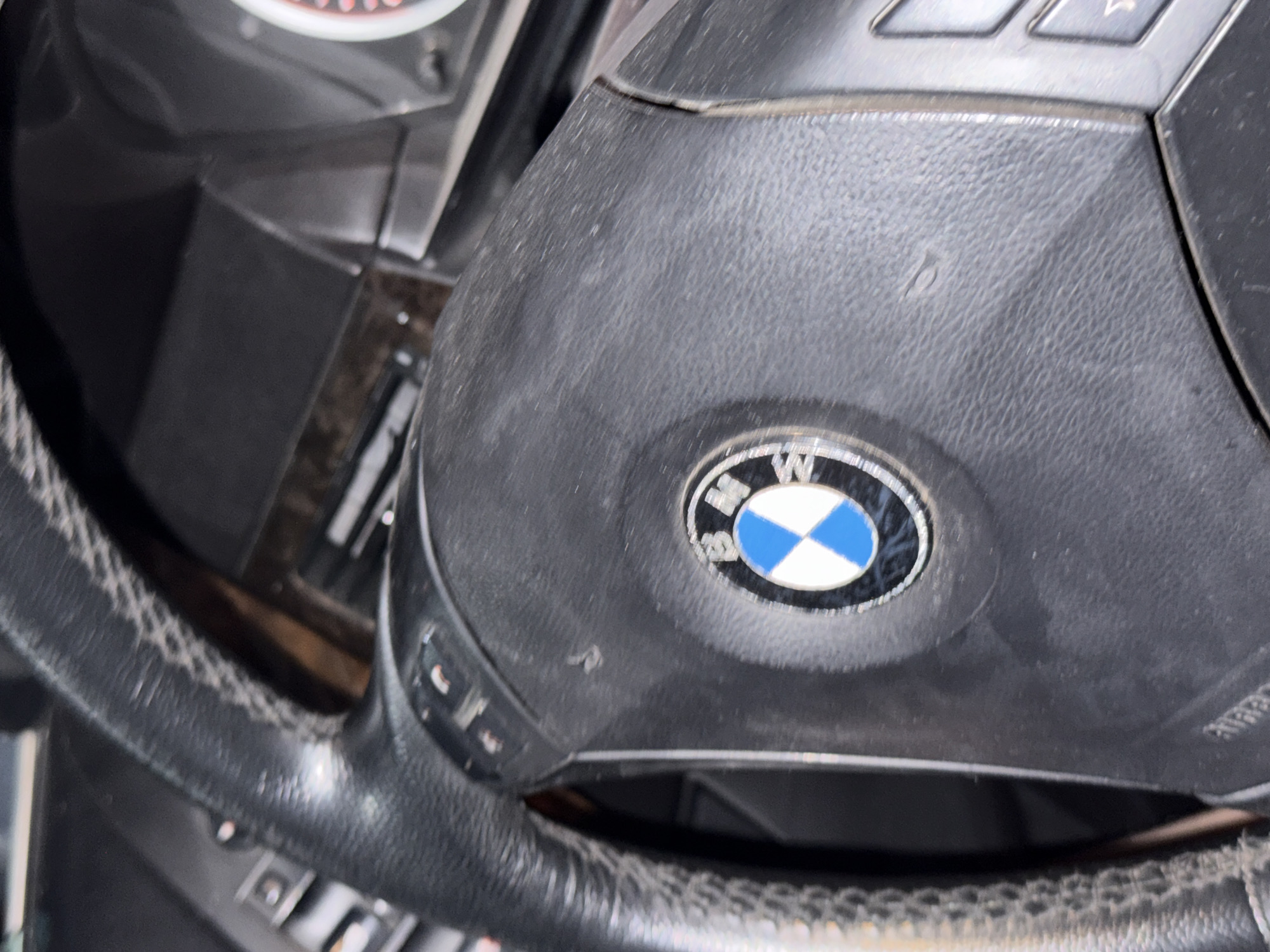 The image shows a close-up of a black leather BMW steering wheel. The BMW logo, with blue and white quadrants, is visible in the center of the steering wheel. Several buttons are present on the steering wheel, including what appear to be media controls. The stitching on the steering wheel is a lighter color, possibly white or gray. The dashboard and some wood trim are visible in the background. The image appears to have been taken inside a car. There is no indication of the location being in Stara Zagora, Bulgaria.
