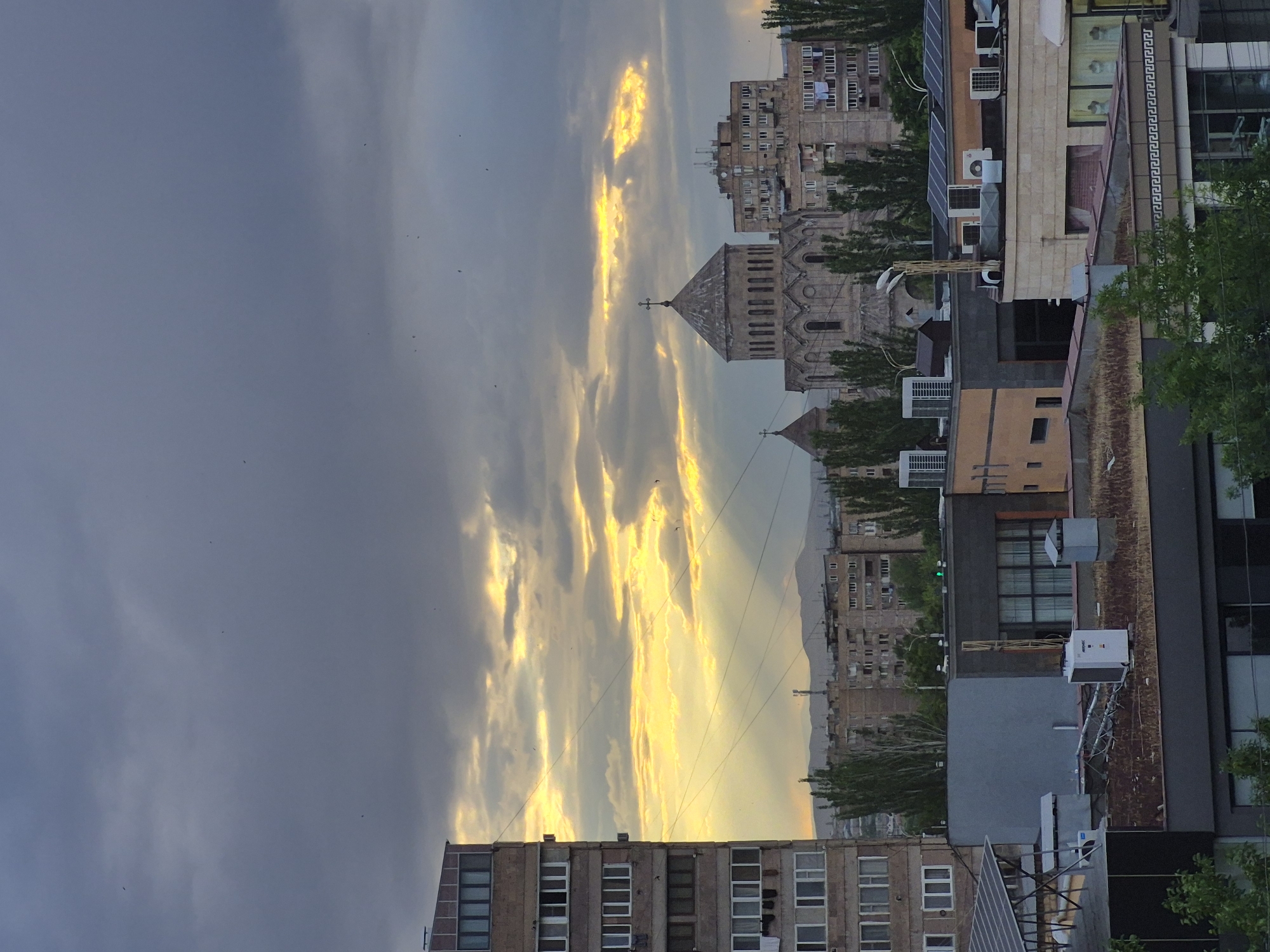 This vertical image depicts an urban landscape in Yerevan, Armenia, bathed in the dramatic light of either sunrise or sunset. The sky dominates the left side, showcasing a striking contrast between dark, bluish-gray clouds and brilliant golden light breaking through, creating visible sunbeams and highlighting the cloud formations. Numerous small, dark specks, likely birds, are visible against the sky.
In the mid-ground, a prominent stone church with a distinct dome and a cross on its spire stands out, flanked by various multi-story buildings. These buildings display diverse architectural styles, with some featuring multiple windows, balconies, and clothes hanging to dry. Many rooftops are adorned with air conditioning units and, in one instance, solar panels. Green trees are interspersed throughout the scene, adding natural elements to the cityscape.
In the distance, the faint outlines of mountains or hills are visible, receding into the hazy light. The golden glow suggests the warm light of a twilight hour, casting a serene yet vibrant atmosphere over the city. No discernible text is clearly visible within the image.
