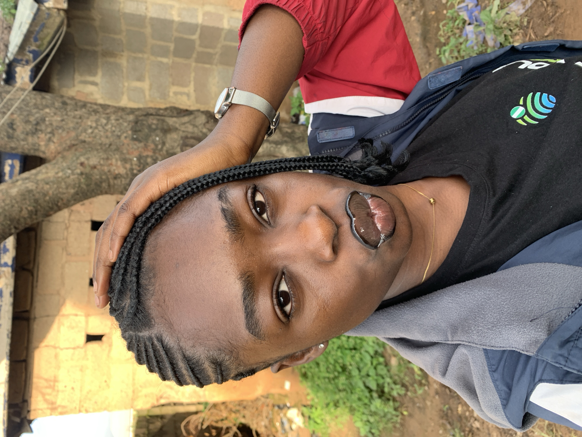 A young Black woman is captured in a medium close-up shot, outdoors, in what appears to be a rural setting. Her right hand is raised, touching her head, with her fingers splayed across her braided hair. She has on a dark t-shirt with a colorful circular logo on the left chest, and a blue and gray hooded jacket with red sleeves. A thin gold chain with a small spherical pendant is visible around her neck. She is wearing a silver mesh-strap watch on her left wrist. Her lips are painted with dark lipstick, creating a distinctive outline around her mouth, with some gloss evident. She is looking directly at the camera. The background includes a textured wall of a building constructed with pale, rough-hewn blocks, a portion of a tree trunk with rough bark, and some greenery and scattered debris. The lighting suggests it is daytime, with shadows indicating the sun's position. The location is identified as Rafin Par, Nigeria.