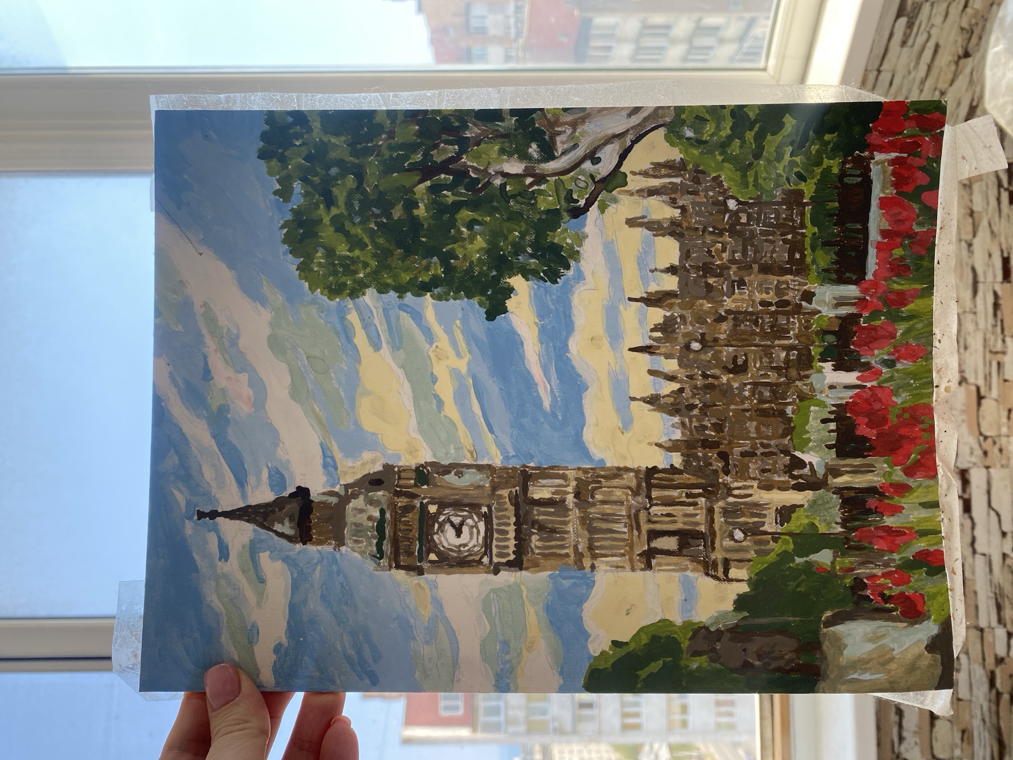 The image is a painting of the Elizabeth Tower, also known as Big Ben, in London, United Kingdom. The painting depicts the iconic clock tower with its surrounding buildings and greenery. In the foreground, there are red flowers, possibly tulips, and a tree on the right side of the painting. The sky is depicted with clouds, suggesting a daytime scene. The painting is held up by a hand in the upper left corner, and it appears to be taped to a surface. The background shows a window with a view of buildings outside, which may be a residential area. The question asks for the location in the city of Chornomorsk, Ukraine, but this is a painting of a landmark in London, not Chornomorsk. There are no people depicted in the painting.
