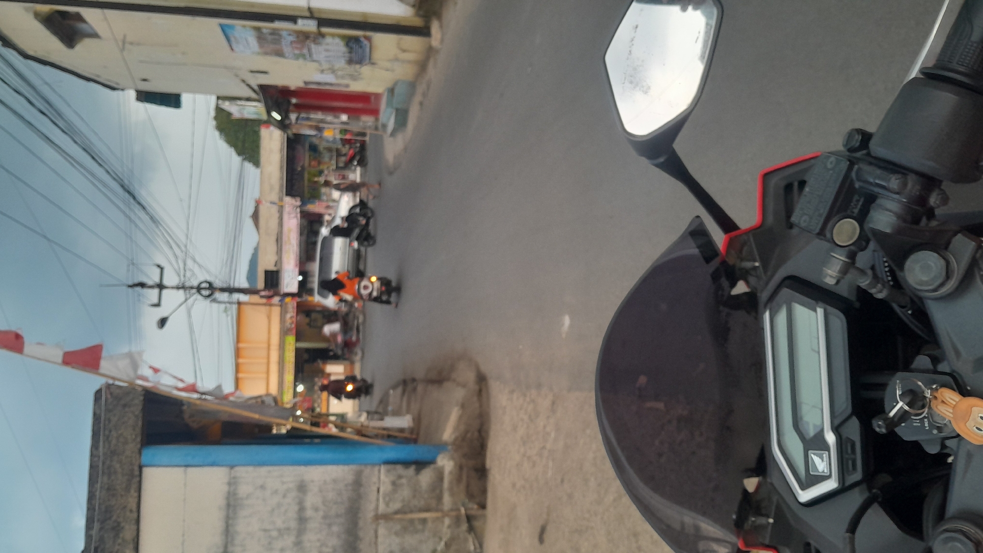 The image depicts a view from the perspective of someone riding a motorcycle on a street in Subang, Indonesia. The street is lined with buildings on both sides, some appearing to be shops or residences. There are several motorcycles on the road, with people riding them. One person in particular is wearing an orange shirt and riding a motorcycle. In the foreground, the handlebars and instrument panel of the motorcycle are visible, indicating the point of view of the rider. Indonesian flags are visible on the left side of the street, suggesting a festive occasion or national pride. The sky is overcast, and the overall lighting suggests it is either early morning or late afternoon. The environment appears to be a typical urban or semi-urban street scene.