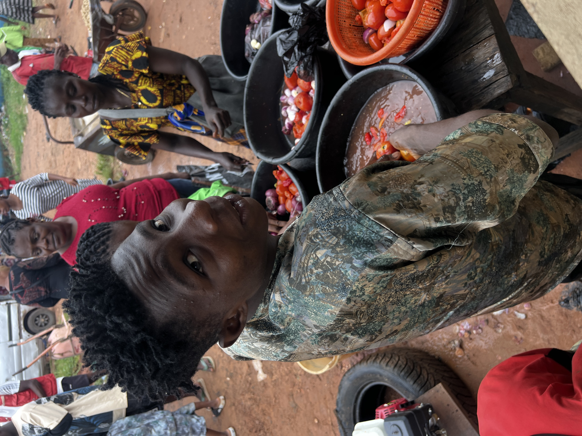 The image depicts a bustling market scene in Nkpor, Nigeria. In the foreground, a young boy with dark, curly hair gazes directly at the camera. He is wearing a camouflage-patterned shirt. Behind him, several women are visible. One woman, wearing a bright red top, is to his left. Further back, another woman in a patterned yellow and black top is looking down. The market appears to be outdoors with a dirt ground. Various goods are displayed in large black basins and an orange plastic basket. These appear to be produce, likely tomatoes and onions, and one basin contains a liquid with peppers. In the background, a white vehicle, possibly a bus, and a wheelbarrow are present, suggesting a busy commercial area. The overall atmosphere is one of active commerce.