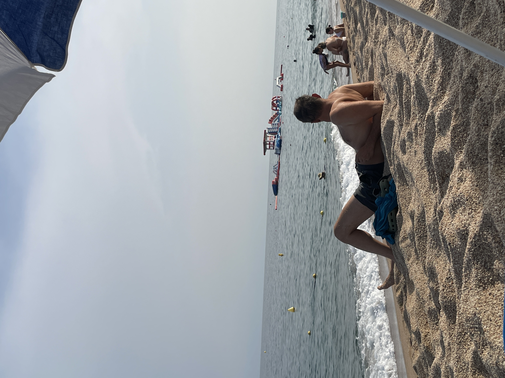 This image shows a beach scene in Canet de Mar, Spain. In the foreground, a man is lying on the sand, facing away from the camera, with his legs extended towards the water. He is wearing dark swim trunks and appears to be relaxing. To his left, a blue towel and a green sandal are placed on the sand near the water's edge.
The sea is calm with small waves lapping at the shore. Several yellow buoys are visible in the water, marking a swimming area. In the middle ground, there is a large inflatable water park structure with slides and platforms. Further out in the sea, several people are visible swimming and playing.
On the beach to the right of the man, a woman and two children are sitting. The woman is wearing a bikini, and one child is standing next to her. Another child is sitting beside her. A pale blue bucket is on the sand near them.
The sky is overcast, with a light gray haze. Part of a white and blue beach umbrella is visible in the upper right corner of the frame. The overall scene depicts a typical summer day at a beach resort.